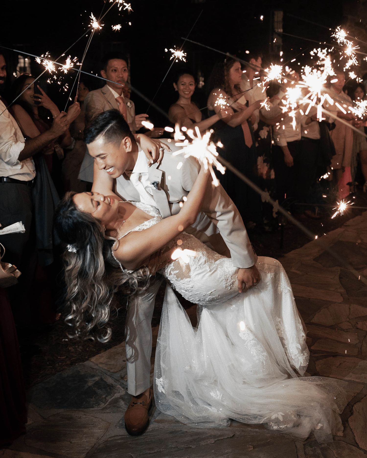 A couple dips and kisses during their sparkler wedding exit surrounded by guests holding sparklers.