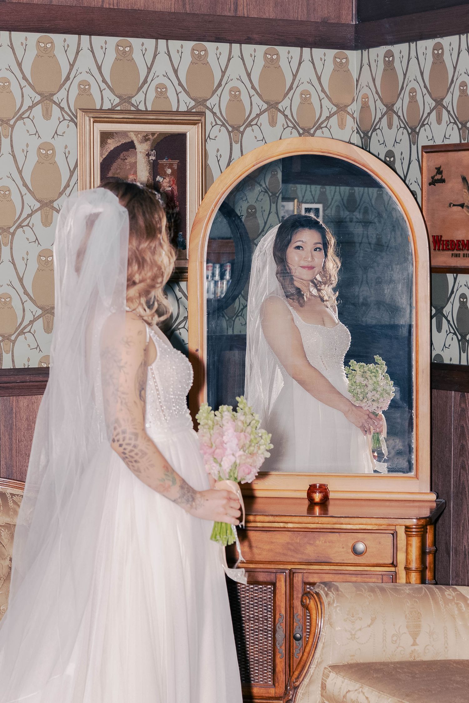 Bride in white gown looking at her reflection in an ornate vintage mirror during wedding preparations.