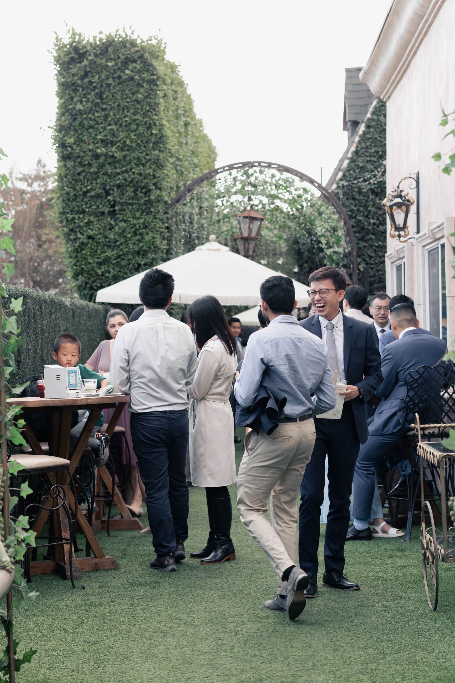 A group of people gather at an outdoor garden party with an arched entryway and green hedges in the background.