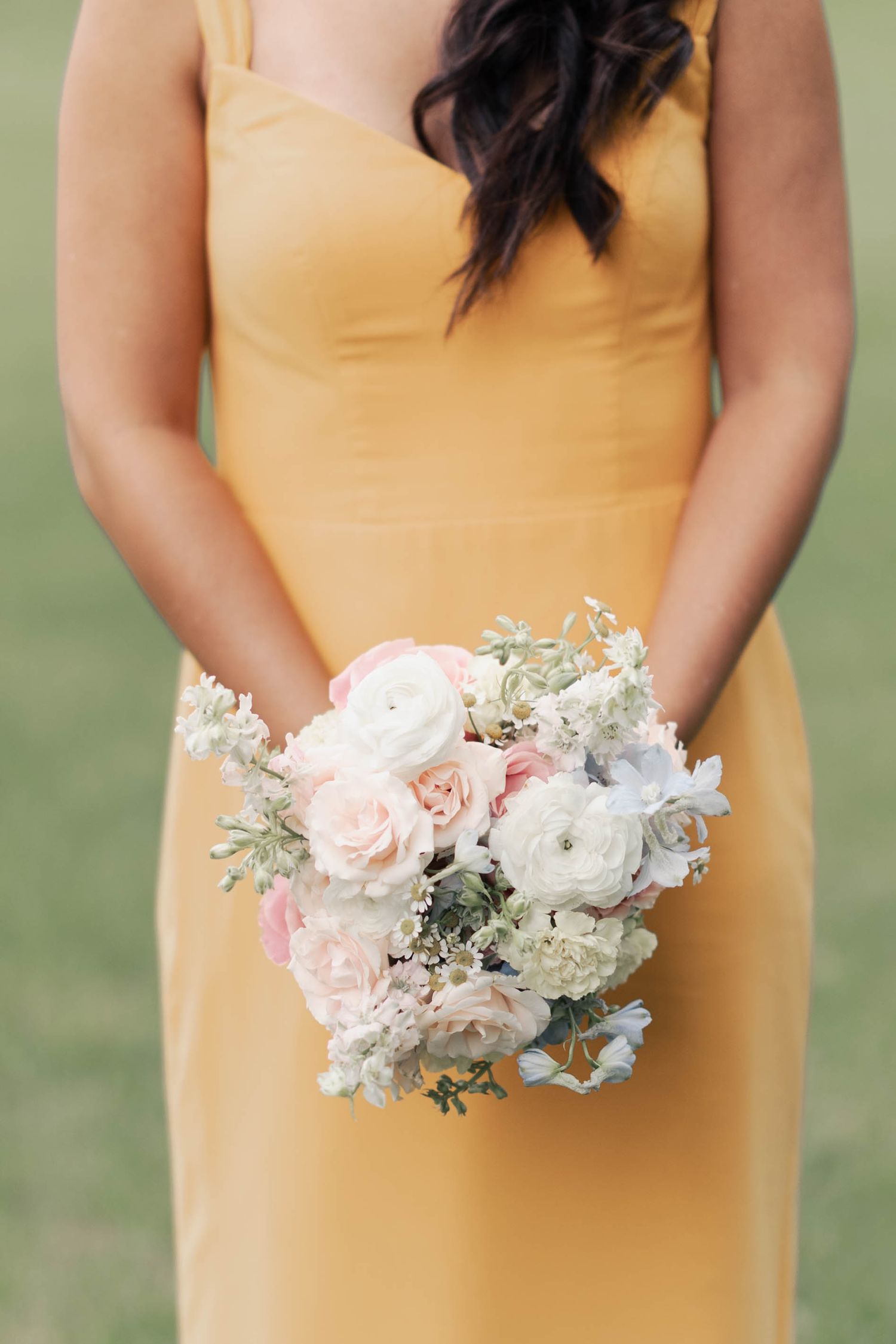 Someone in a yellow bridesmaid dress holds a delicate bouquet of pink and white roses with greenery against a grassy background.