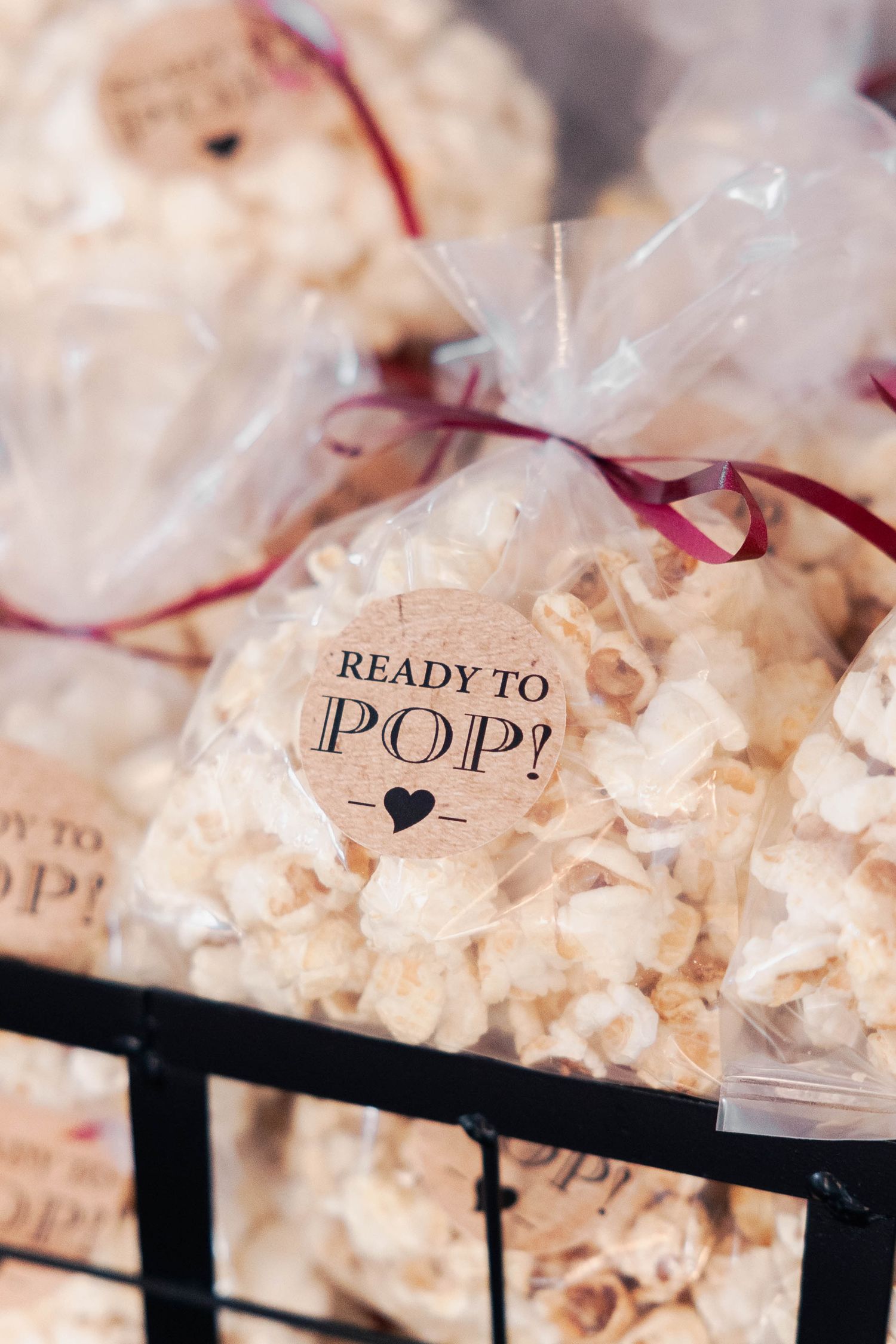 Clear plastic bags filled with popcorn labeled 'Ready to Pop' with burgundy ribbons displayed in a black basket.