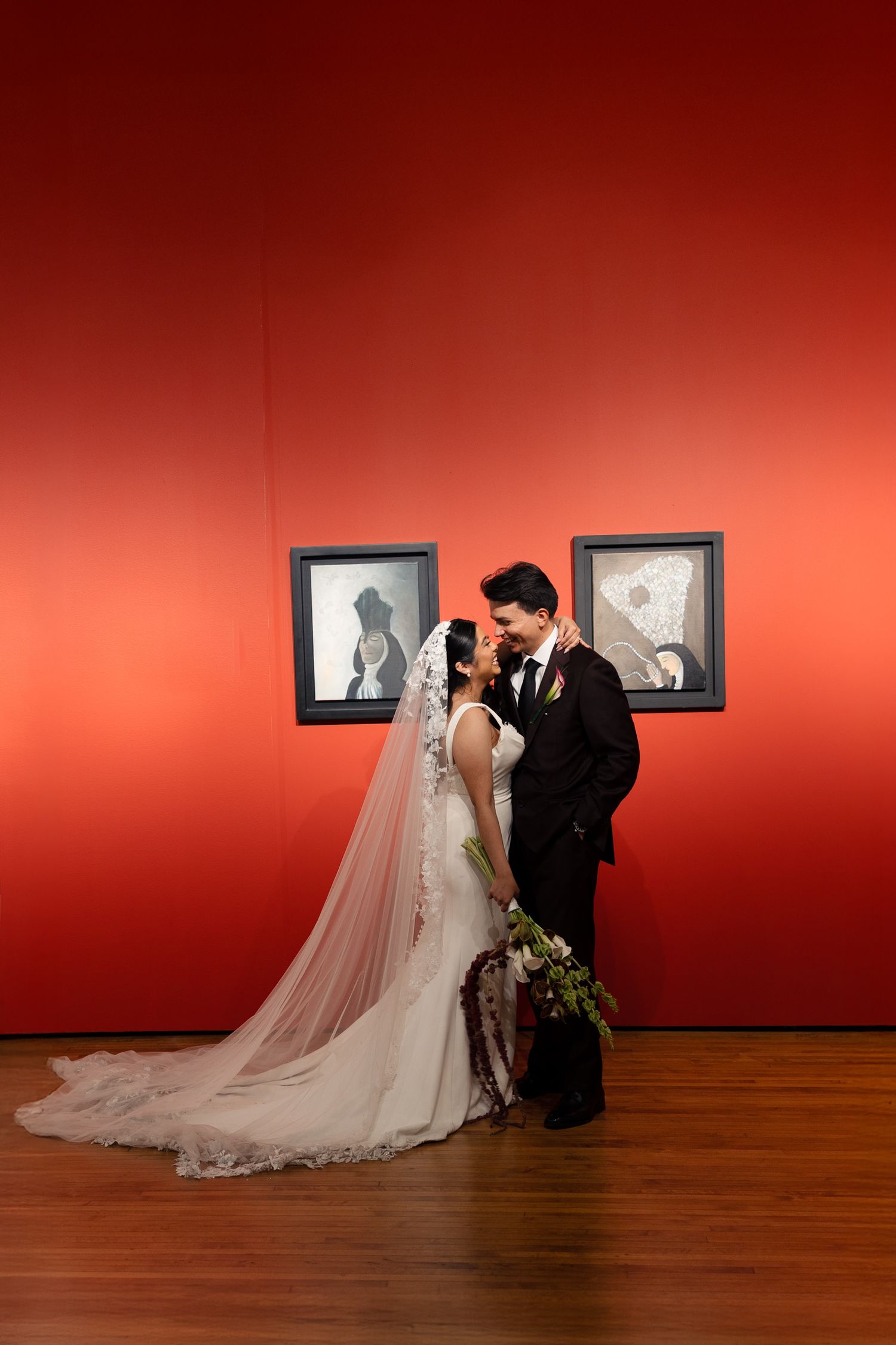 Bride and groom share a romantic moment in a red art gallery, bride wearing a long veil holding a bouquet.