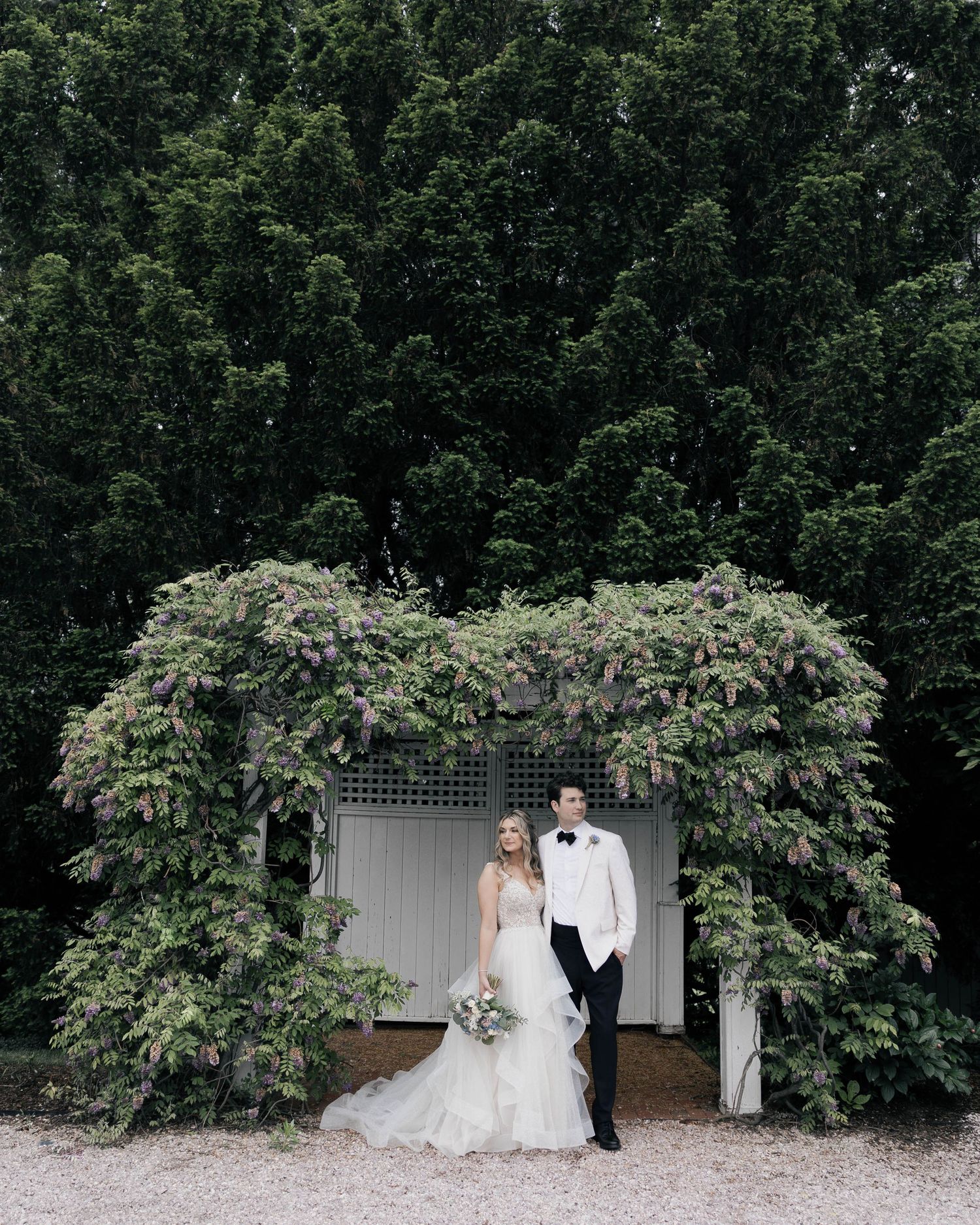 Wedding couple poses in front of a lush garden arbor with greenery and flowers creating a romantic backdrop.