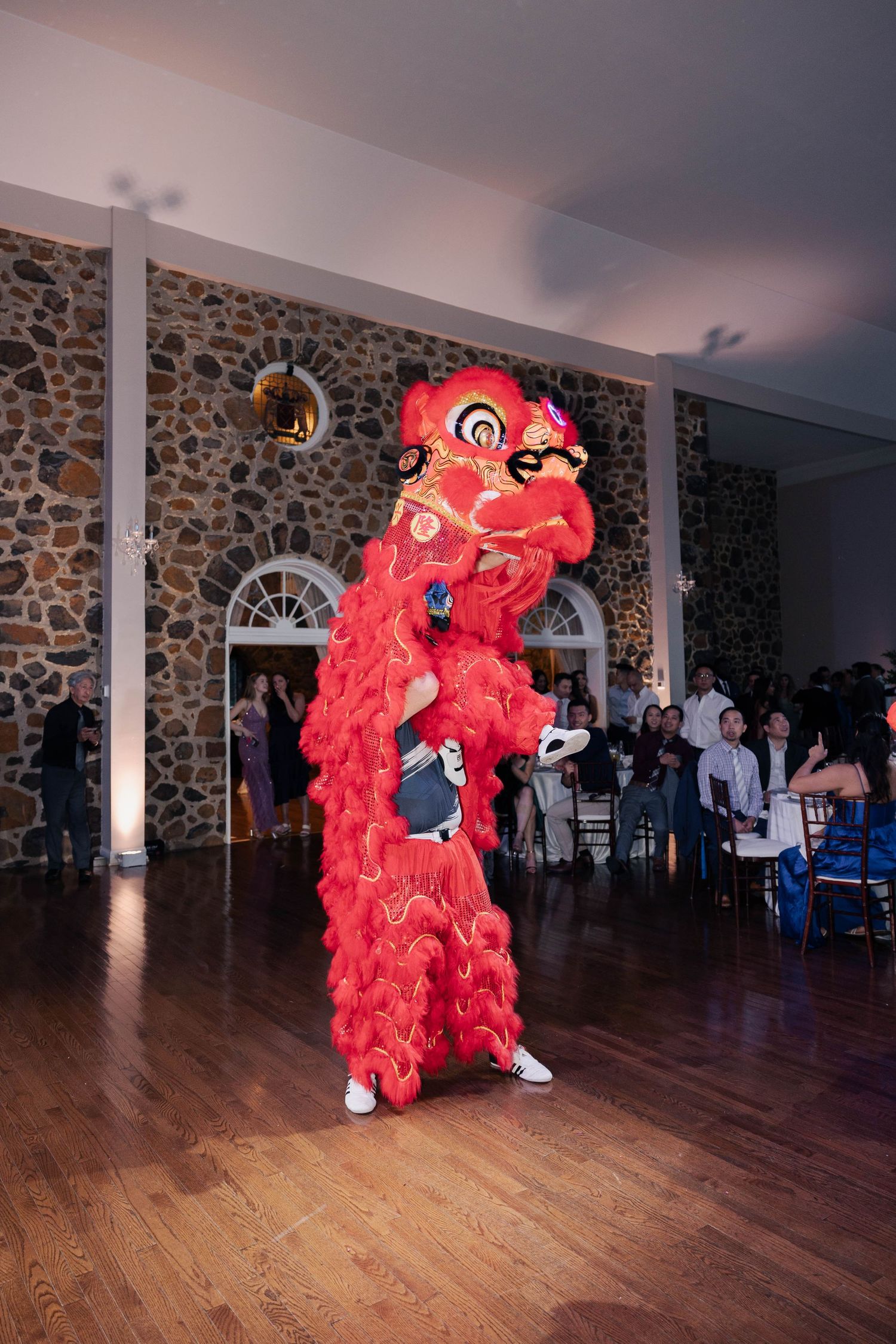 Red lion dancers perform traditional dance moves on hardwood floor at wedding reception.