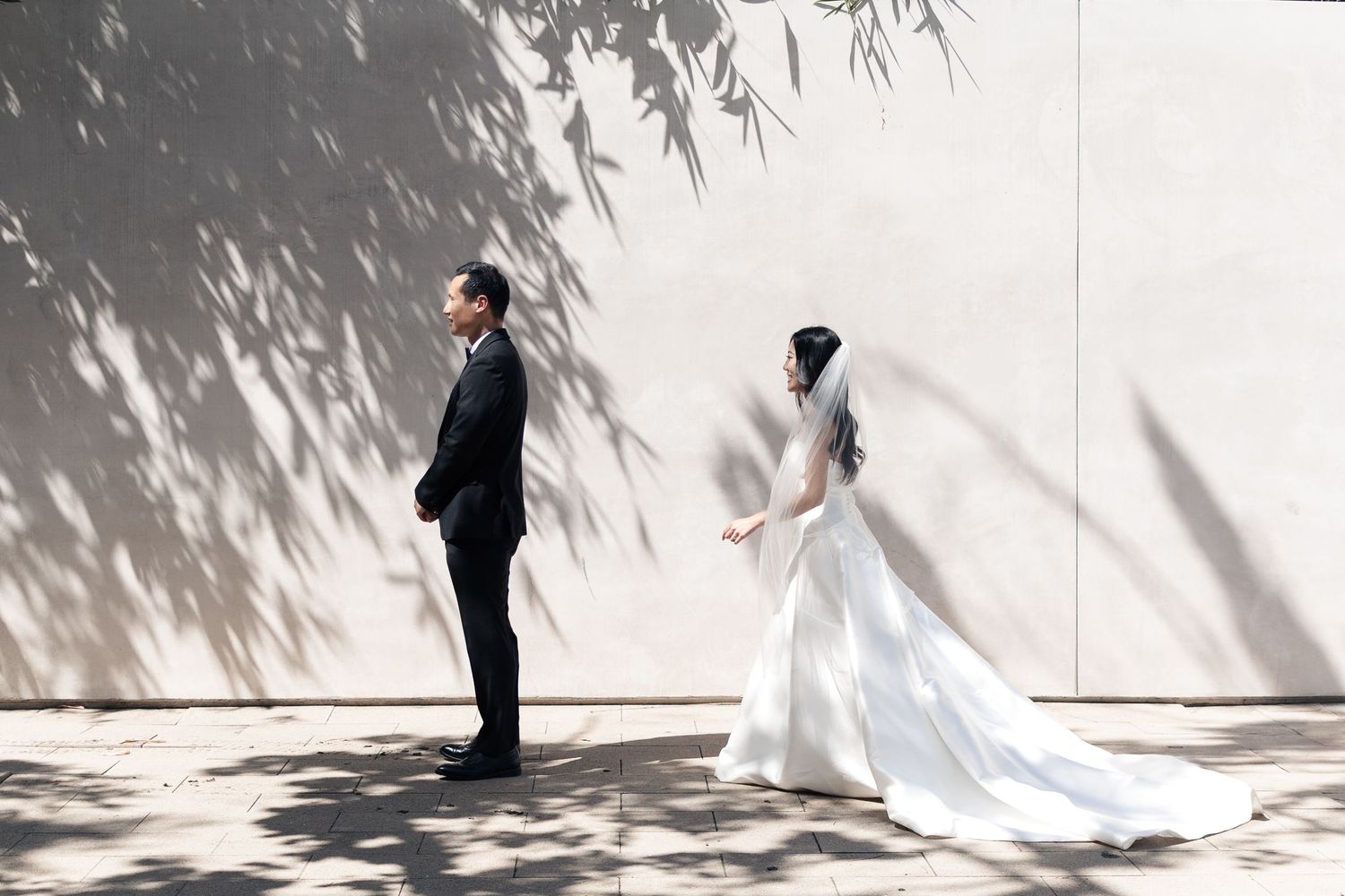 Bride in white gown and groom in black suit share a first look moment against a sunlit wall with bamboo shadows.
