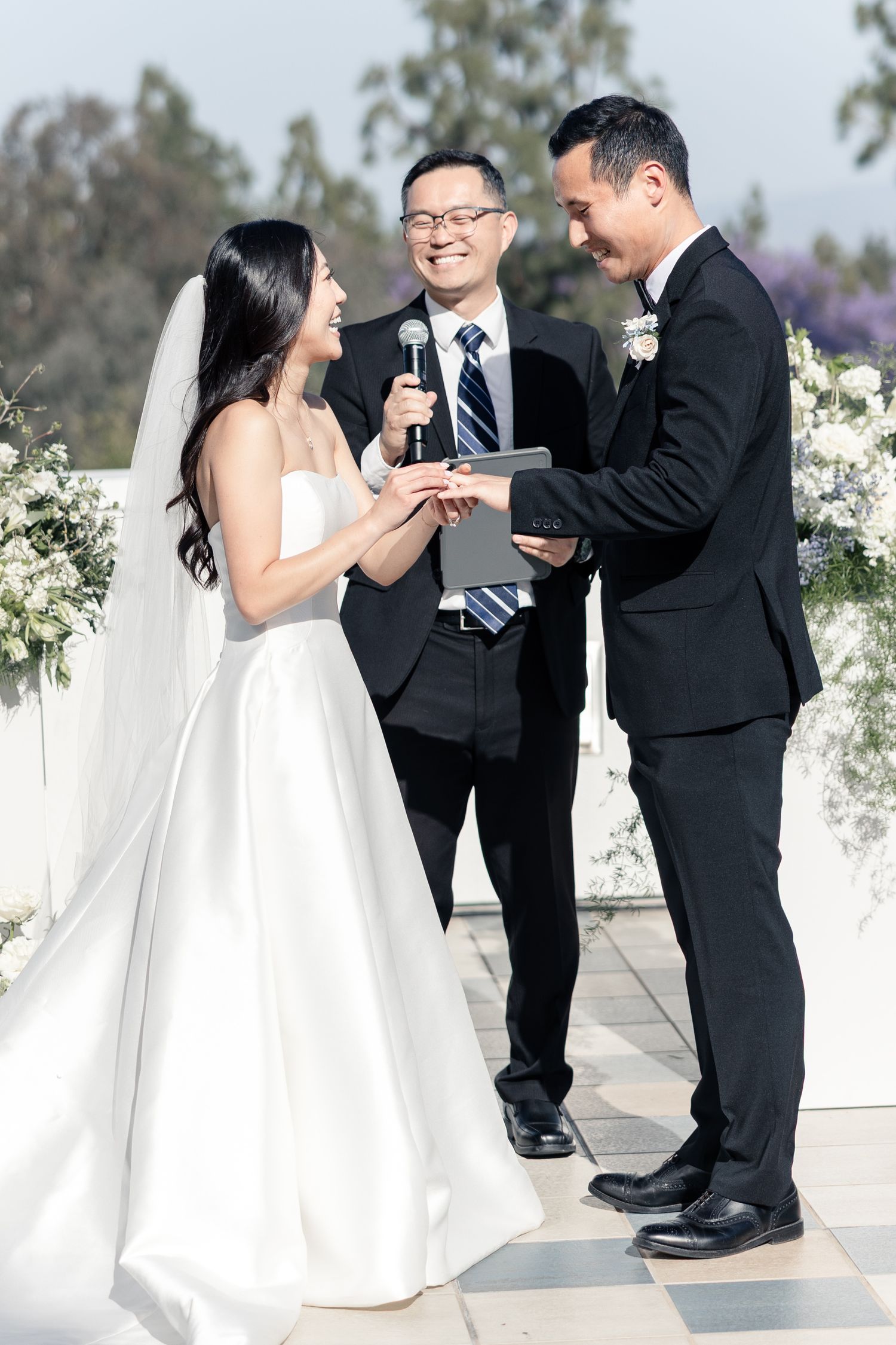 Bride places wedding ring on groom's finger during outdoor ceremony while officiant smiles and holds microphone.