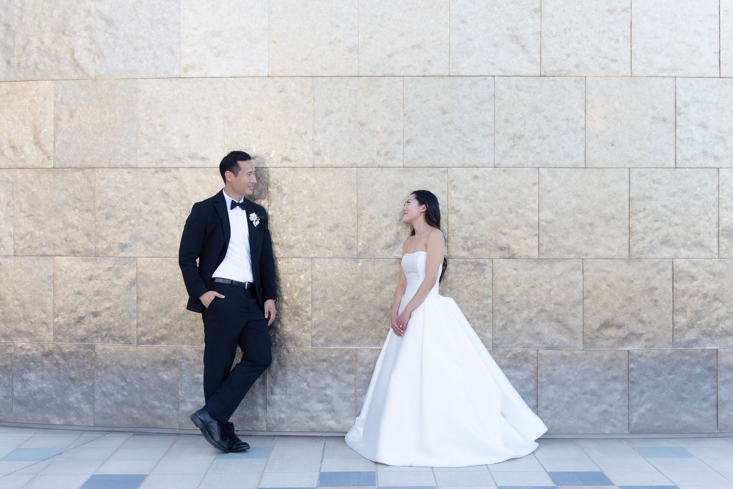 Bride in white ball gown and groom in black tuxedo share a romantic moment against a stone wall.