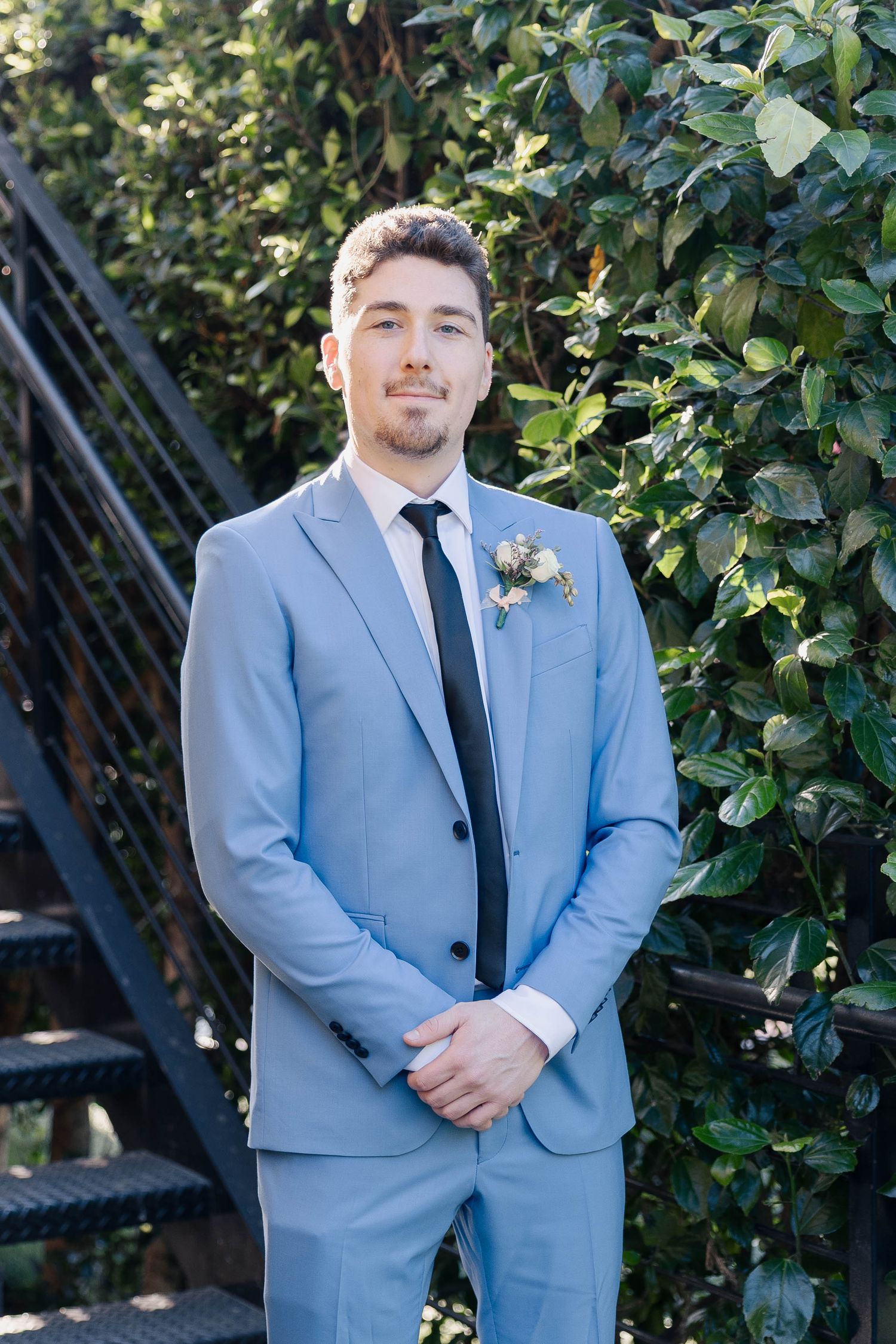 Individual in light blue suit with boutonniere poses against green foliage and metal stairs.
