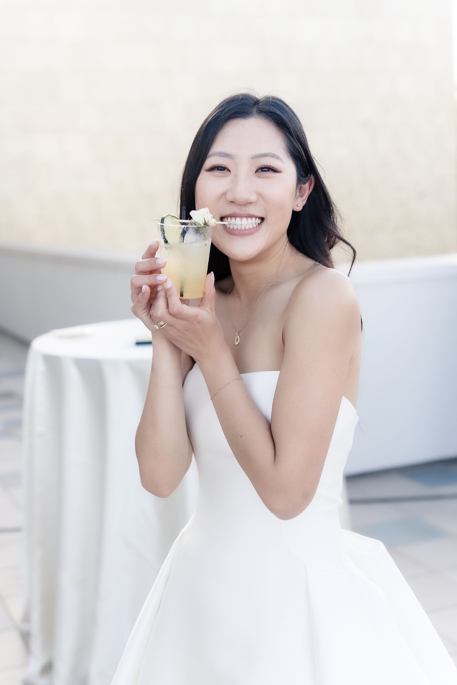 Smiling Asian bride in strapless white gown holding a yellow cocktail drink at an outdoor wedding reception.