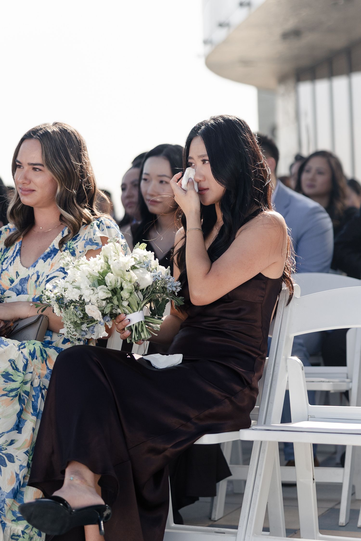 Emotional wedding guest in brown dress wiping tears while holding white bouquet during outdoor ceremony.