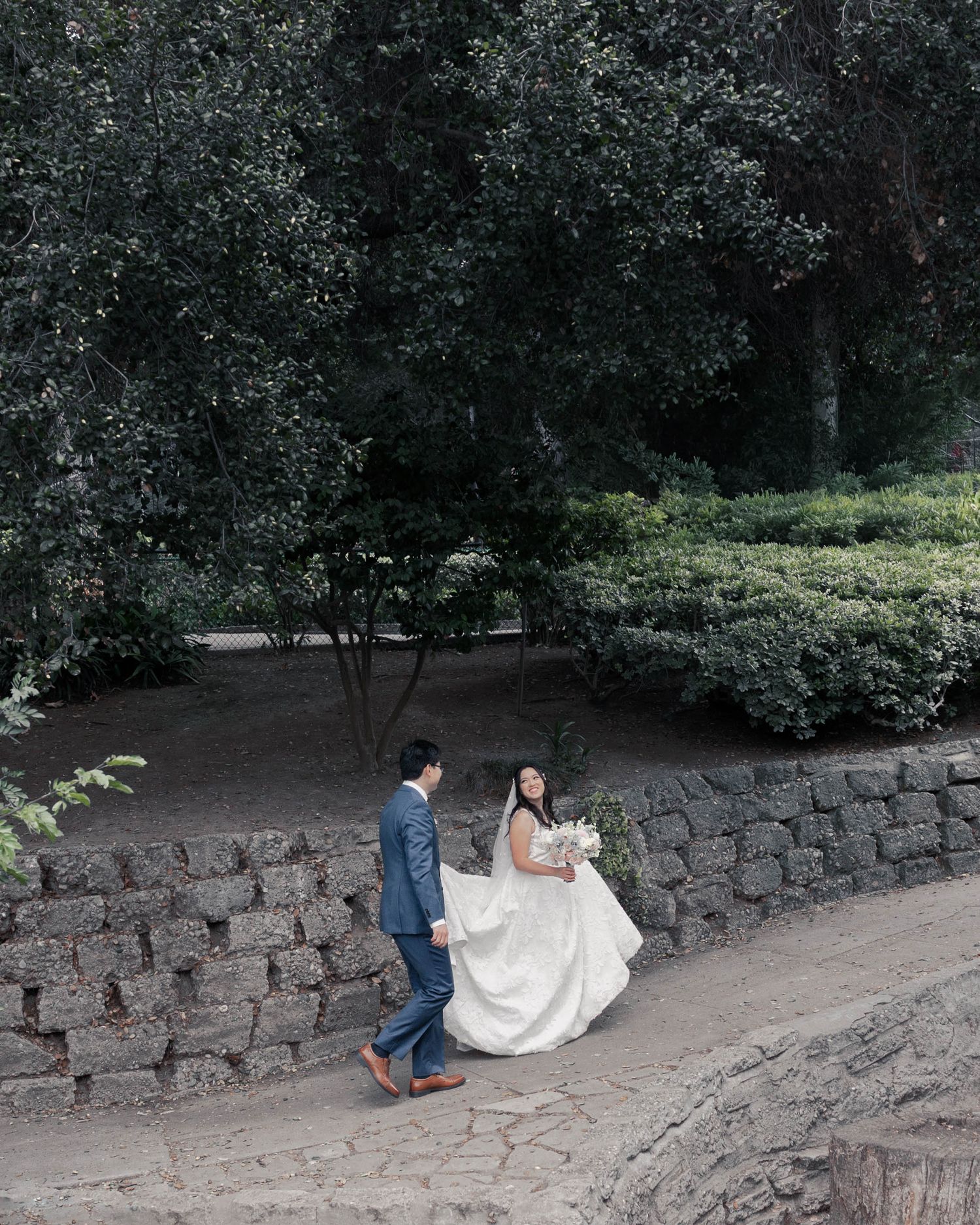 A couple in wedding attire playfully pose on a stone retaining wall in a wooded park setting.