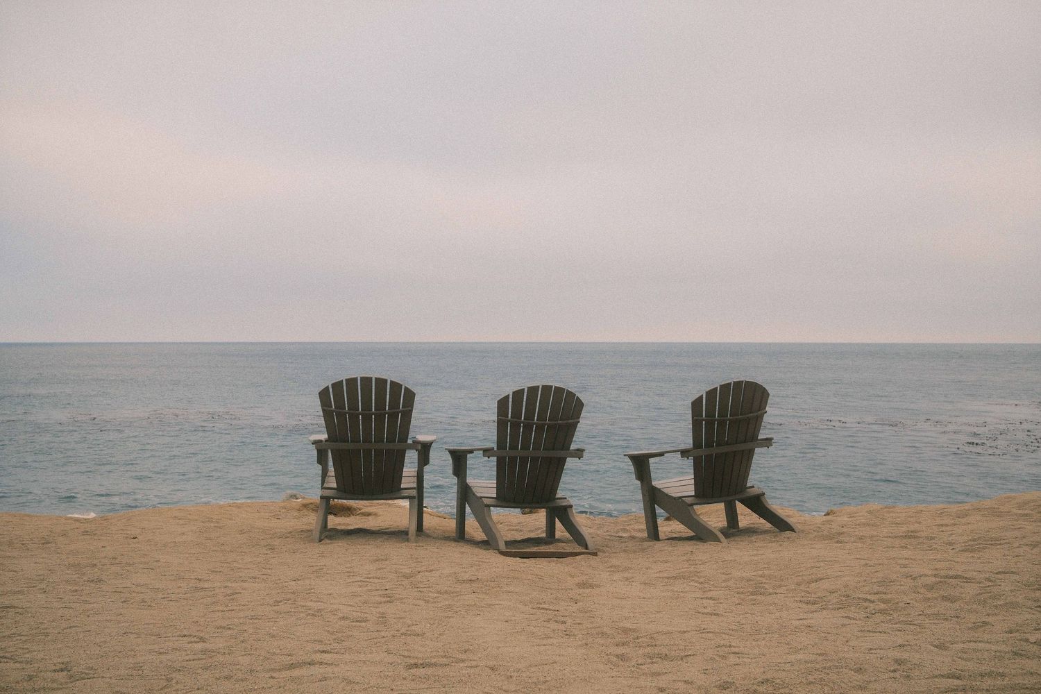 Three empty Adirondack chairs face the ocean at sunset on a sandy beach overlook.