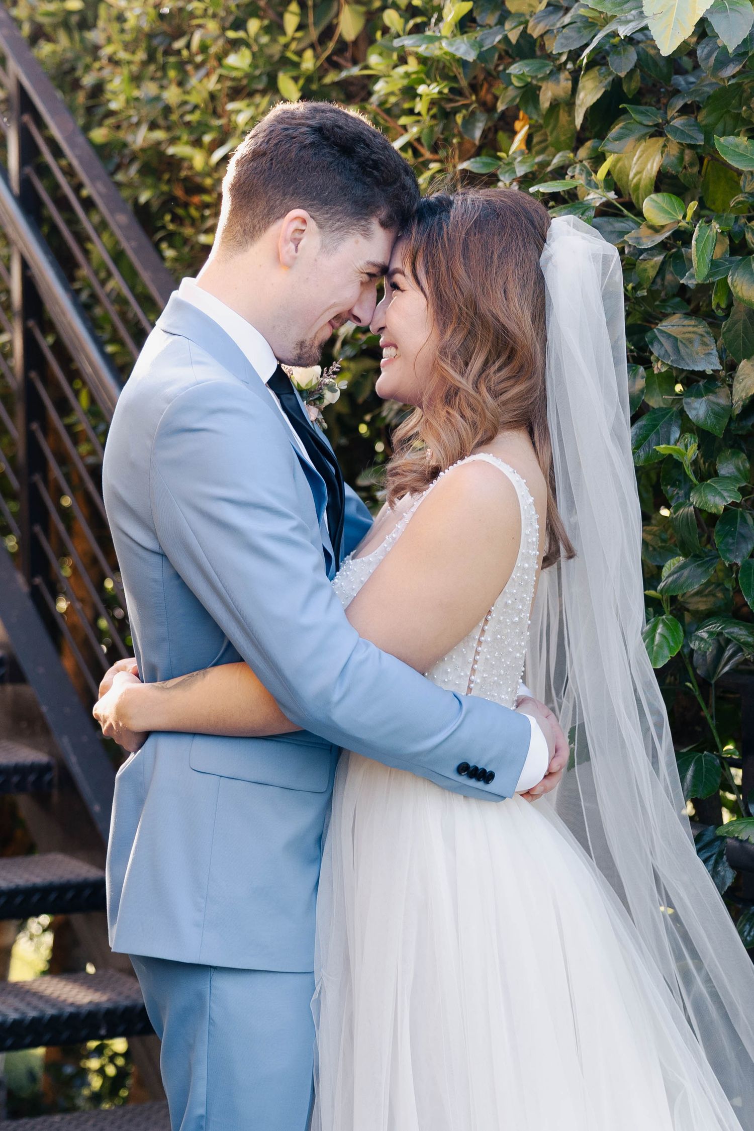 A romantic wedding couple embraces outdoors in front of lush greenery wearing light blue suit and white gown.