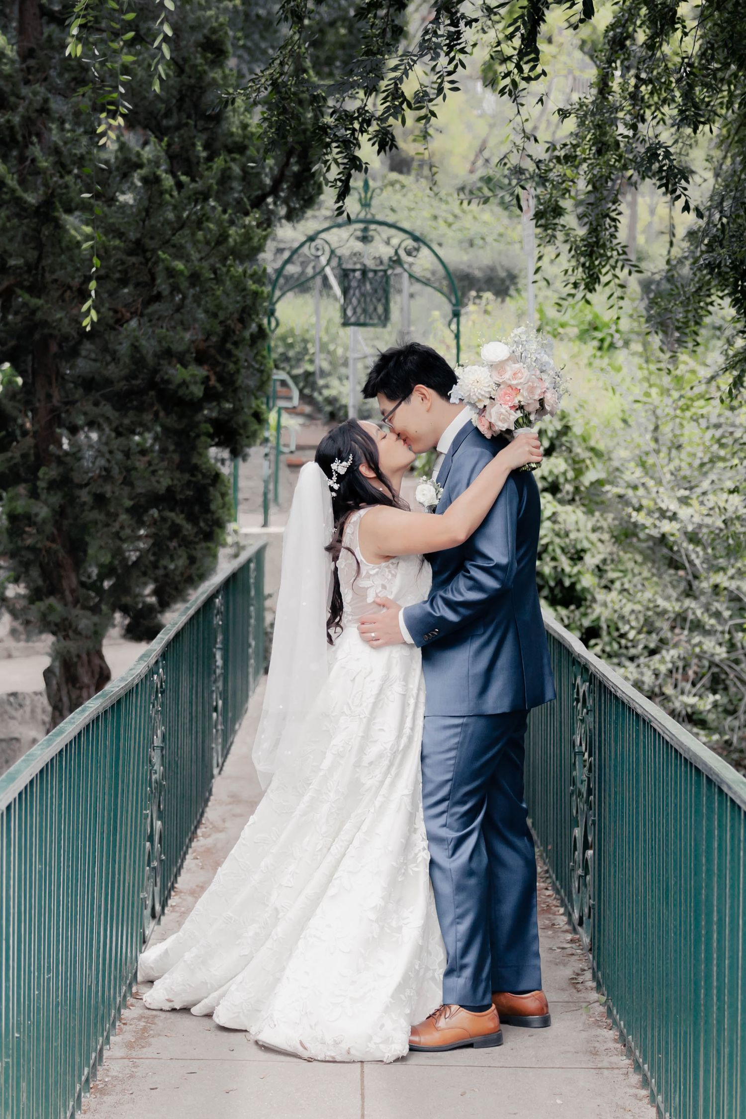 A couple in wedding attire share romantic moments on a pathway with green fencing and trees in the background.