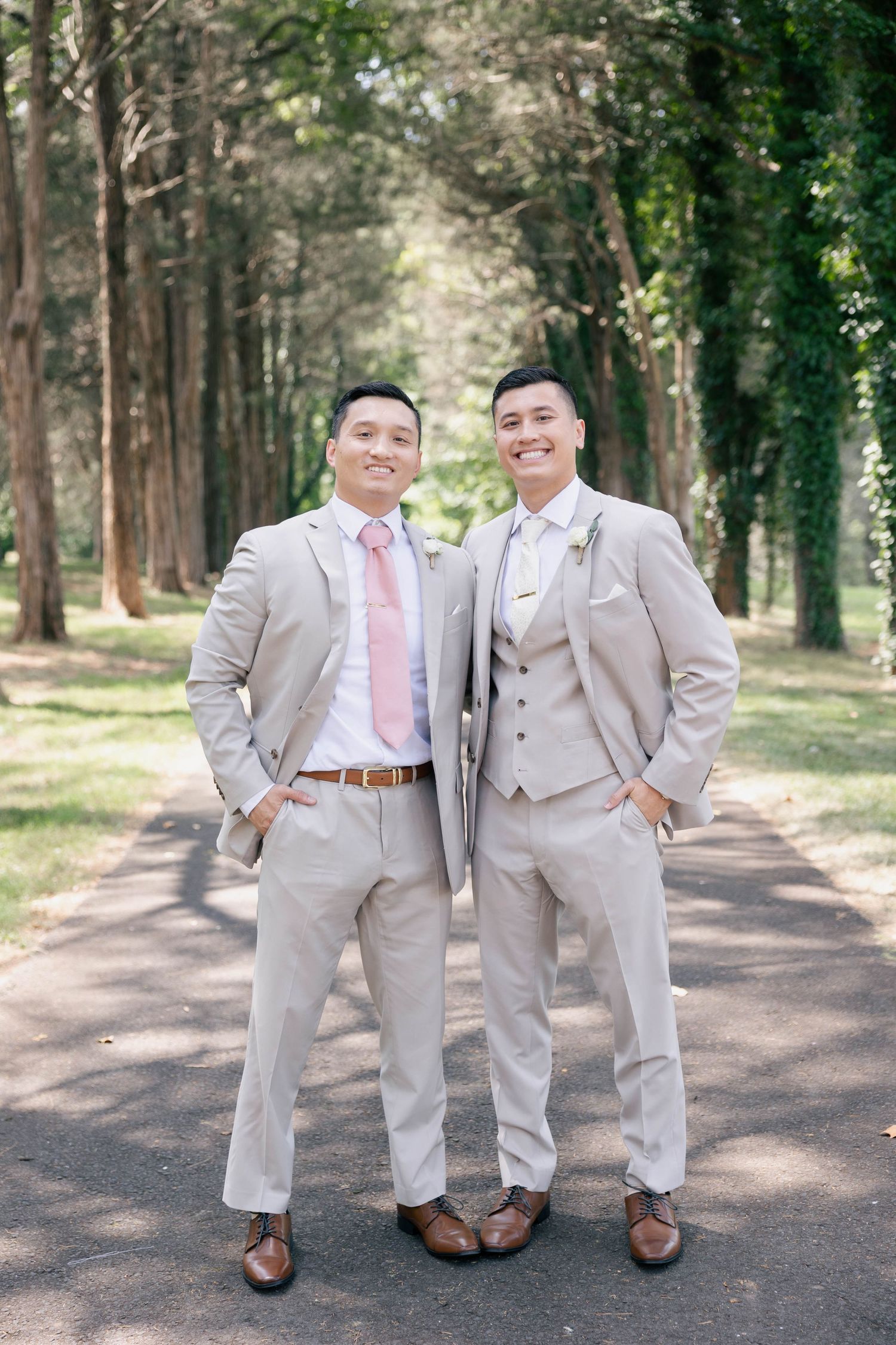 Wedding party members in coordinated gray suits stand casually on wooded path.Groom and groomsmen wedding outfit inspiration in tan suit.