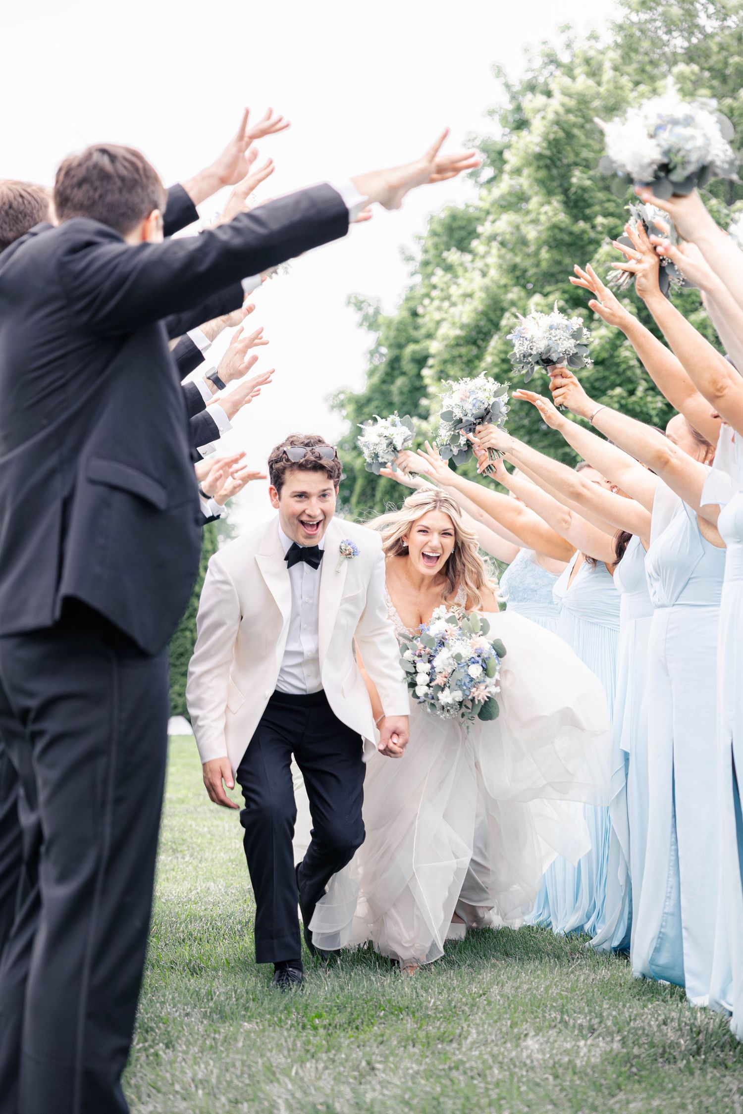 Wedding party in light blue dresses and dark suits jumping joyfully together on grassy lawn during outdoor celebration.