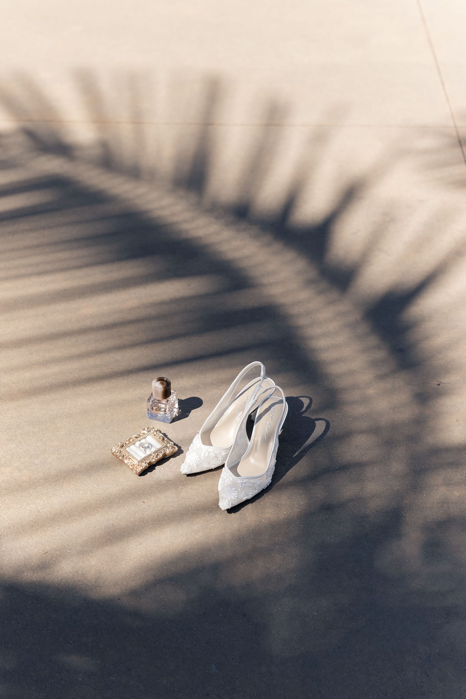 White bridal heels and accessories rest on sandy ground beneath a palm leaf shadow in warm sunlight.