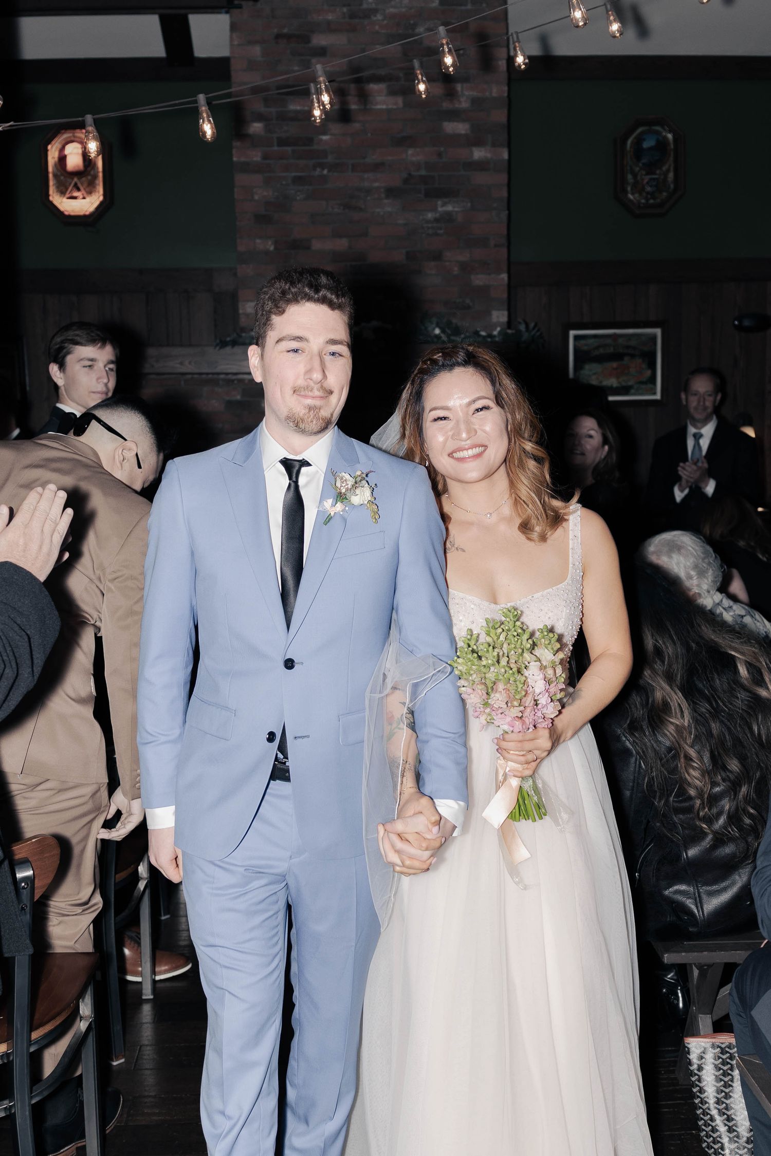Wedding couple in light blue suit and white dress walking through reception venue.