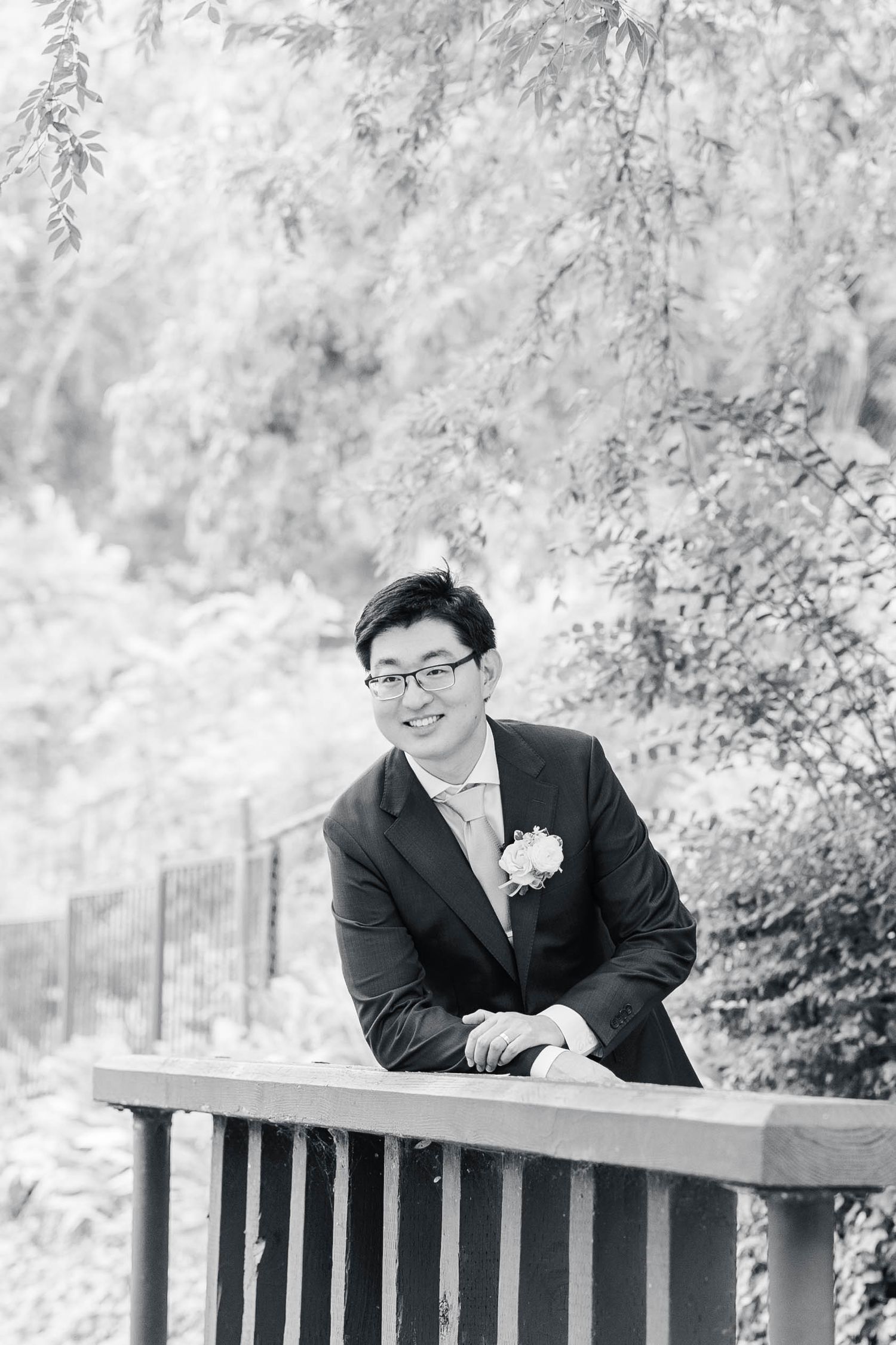 Black and white portrait of someone in formal attire leaning on a wooden railing in a garden setting.