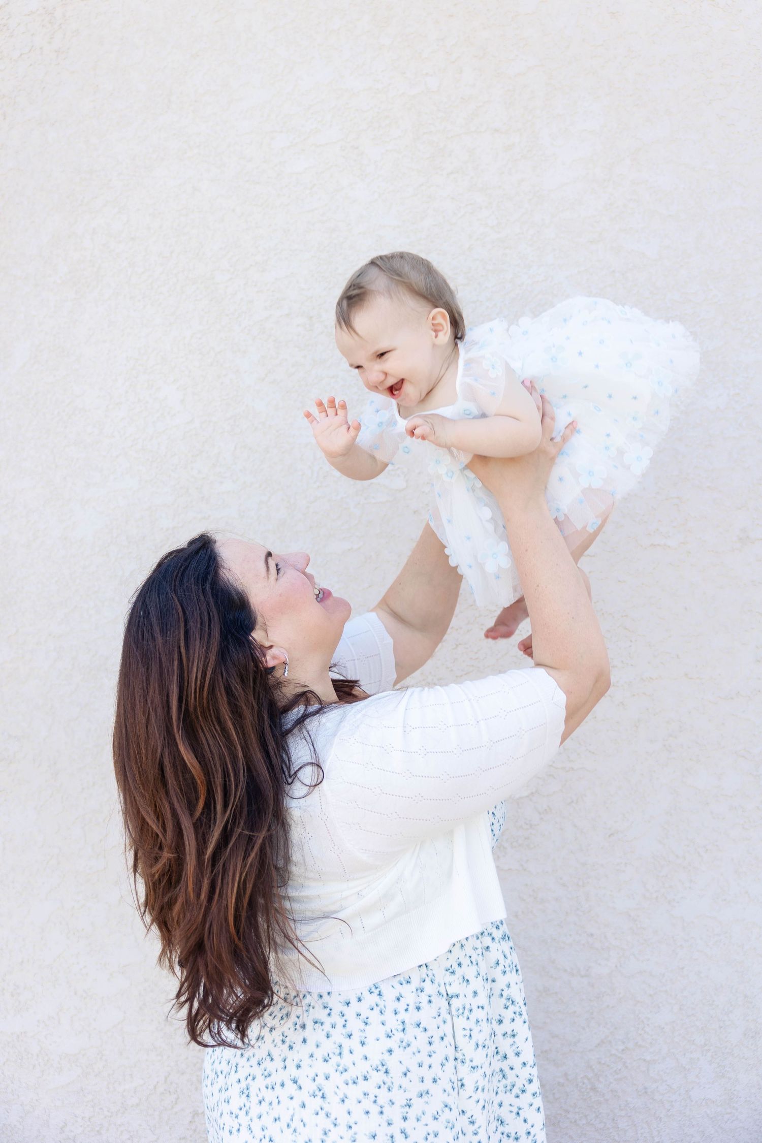 Mother lifting baby in white outfits against light background.