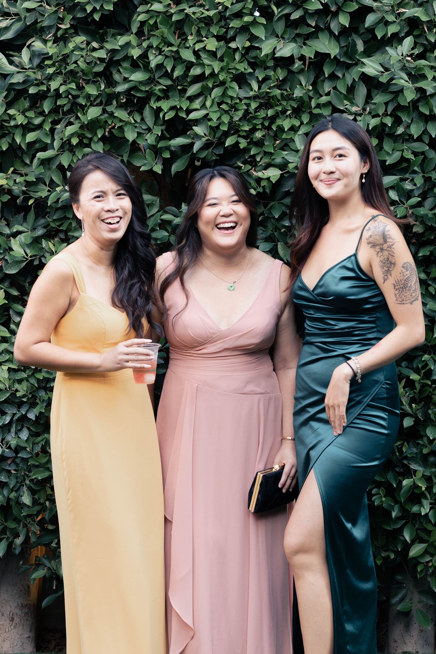 Three women in elegant dresses of yellow, pink and teal stand together laughing against a leafy green backdrop.
