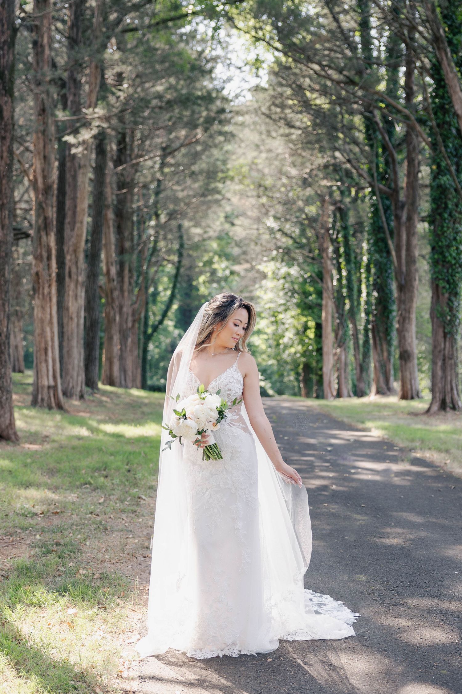 Bride in white lace gown walking down tree-lined path carrying classic white bouquet.