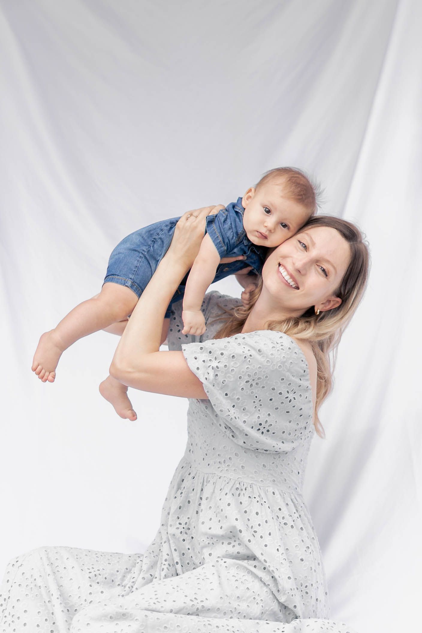 Parent and baby in white dress and denim share joyful moments against white backdrop in studio photoshoot series.