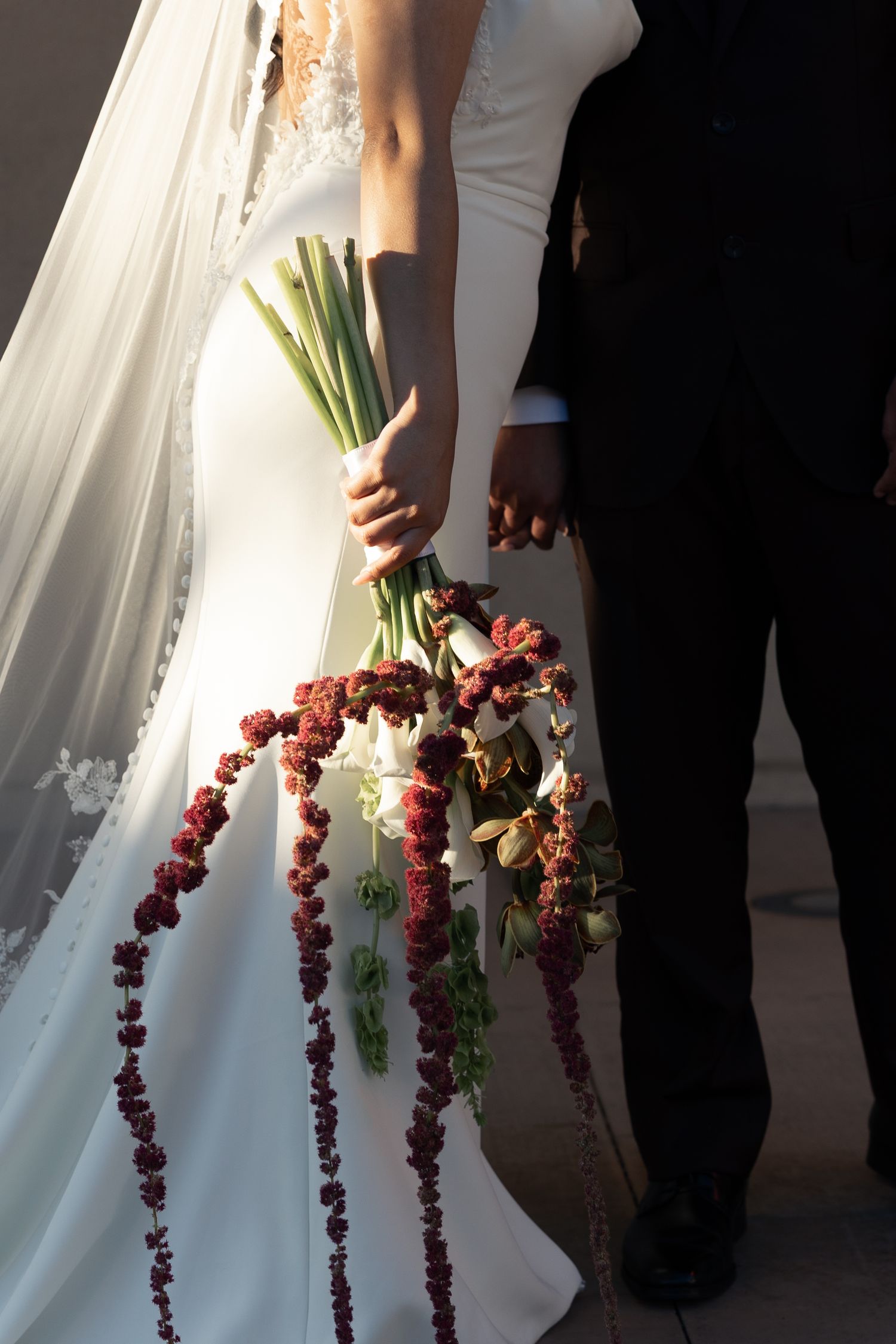 Bride in white lace gown holding cascading bouquet of red amaranthus and white flowers beside groom in black suit.