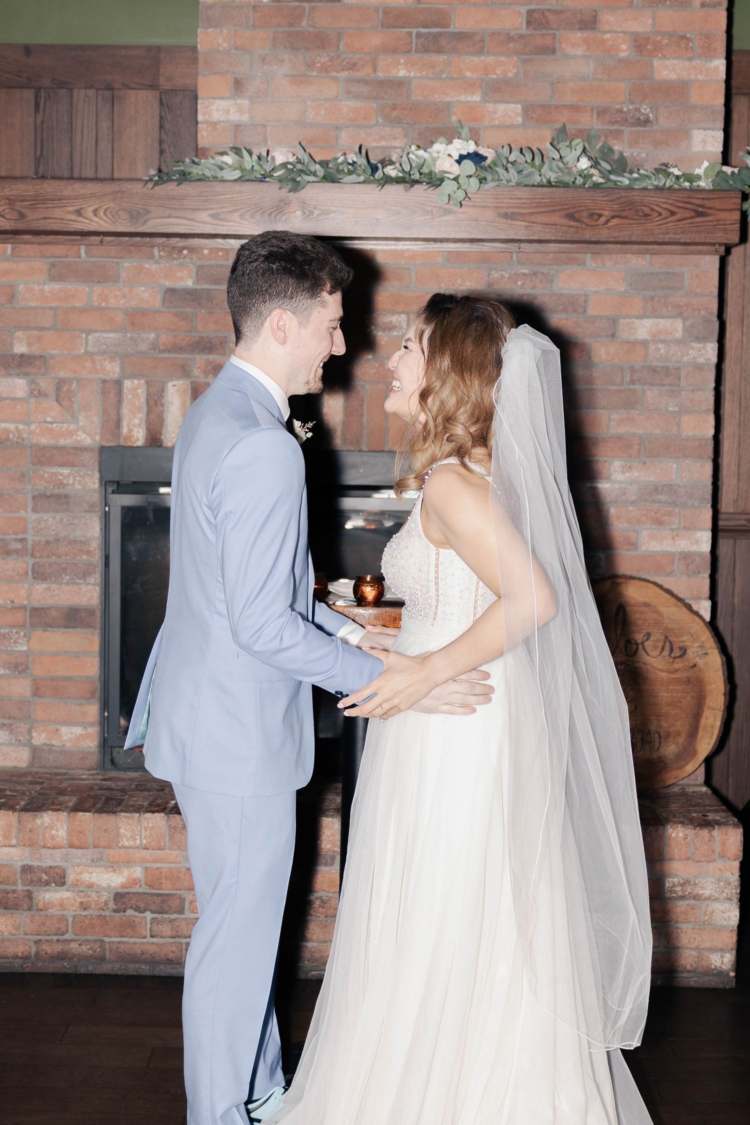 Couple dancing in elegant wedding attire against brick wall backdrop.
