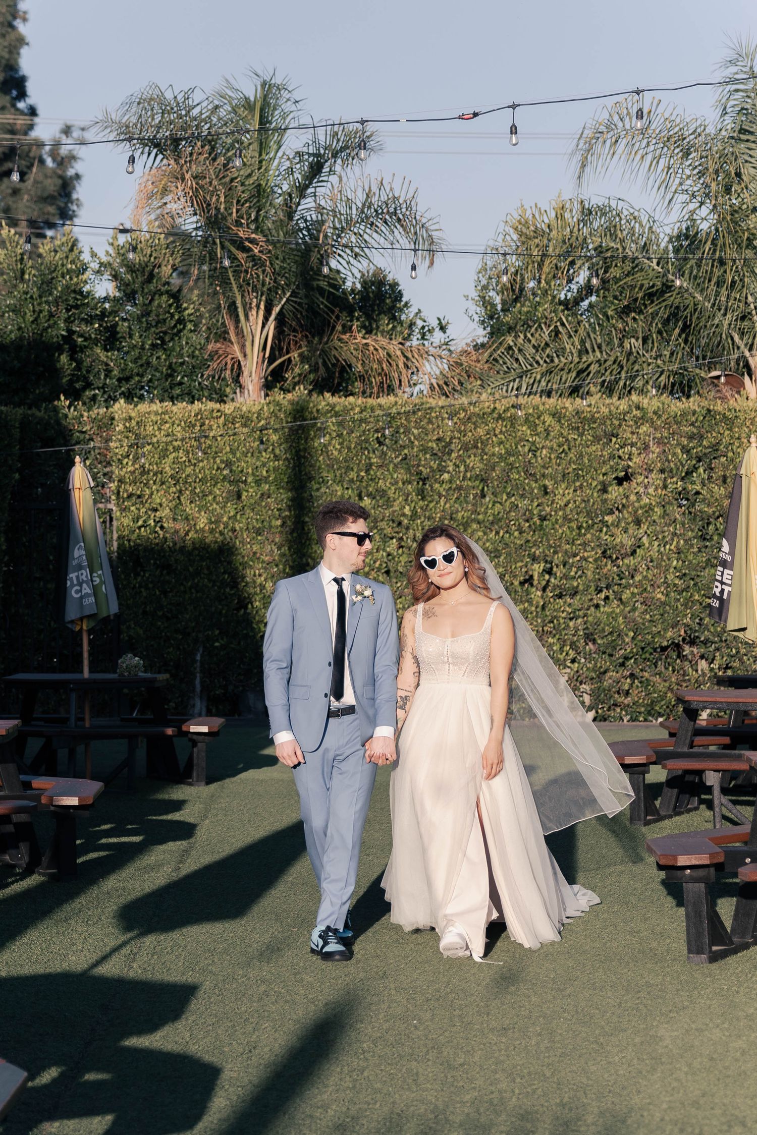 A couple strolls hand in hand across a lawn with string lights overhead and hedges in the background.
