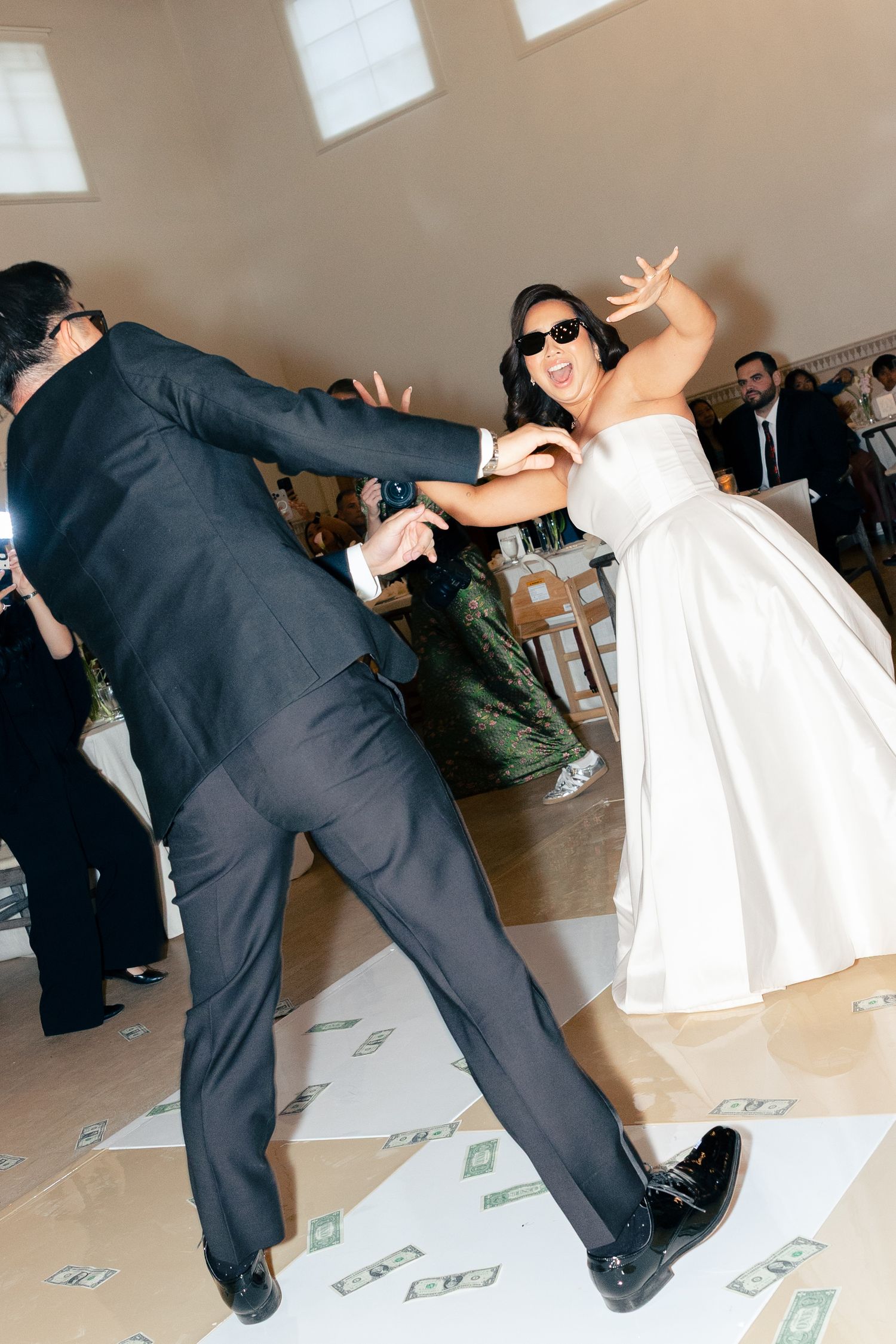Bride in white dress and sunglasses dances energetically with groom in black suit on money-covered dance floor.
