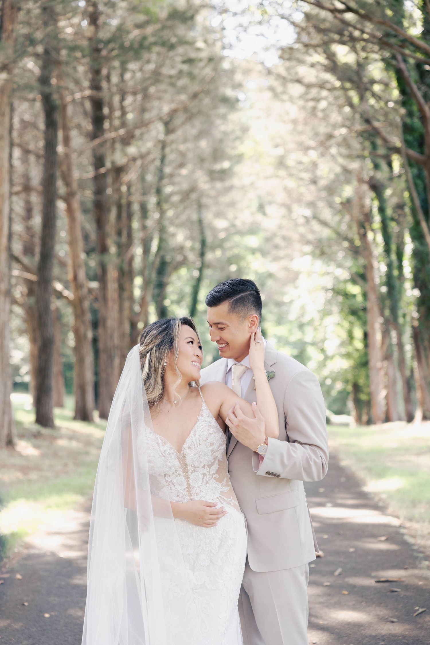 A newlywed couple shares an intimate moment on a tree-lined path in their wedding attire.