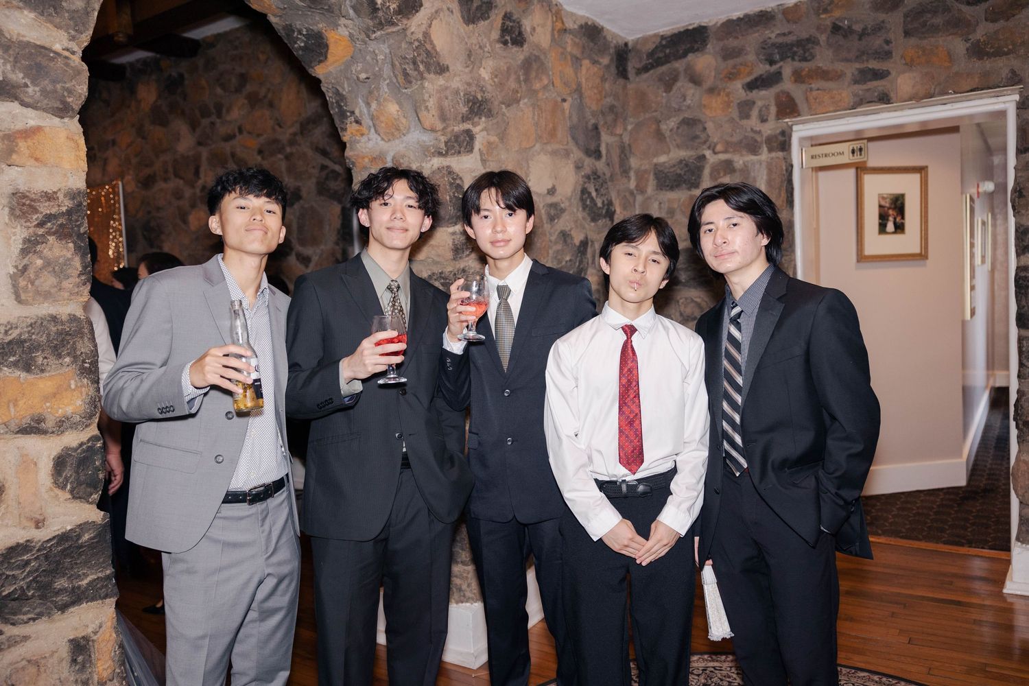 Group of wedding guests in formal suits posing together against stone wall backdrop.