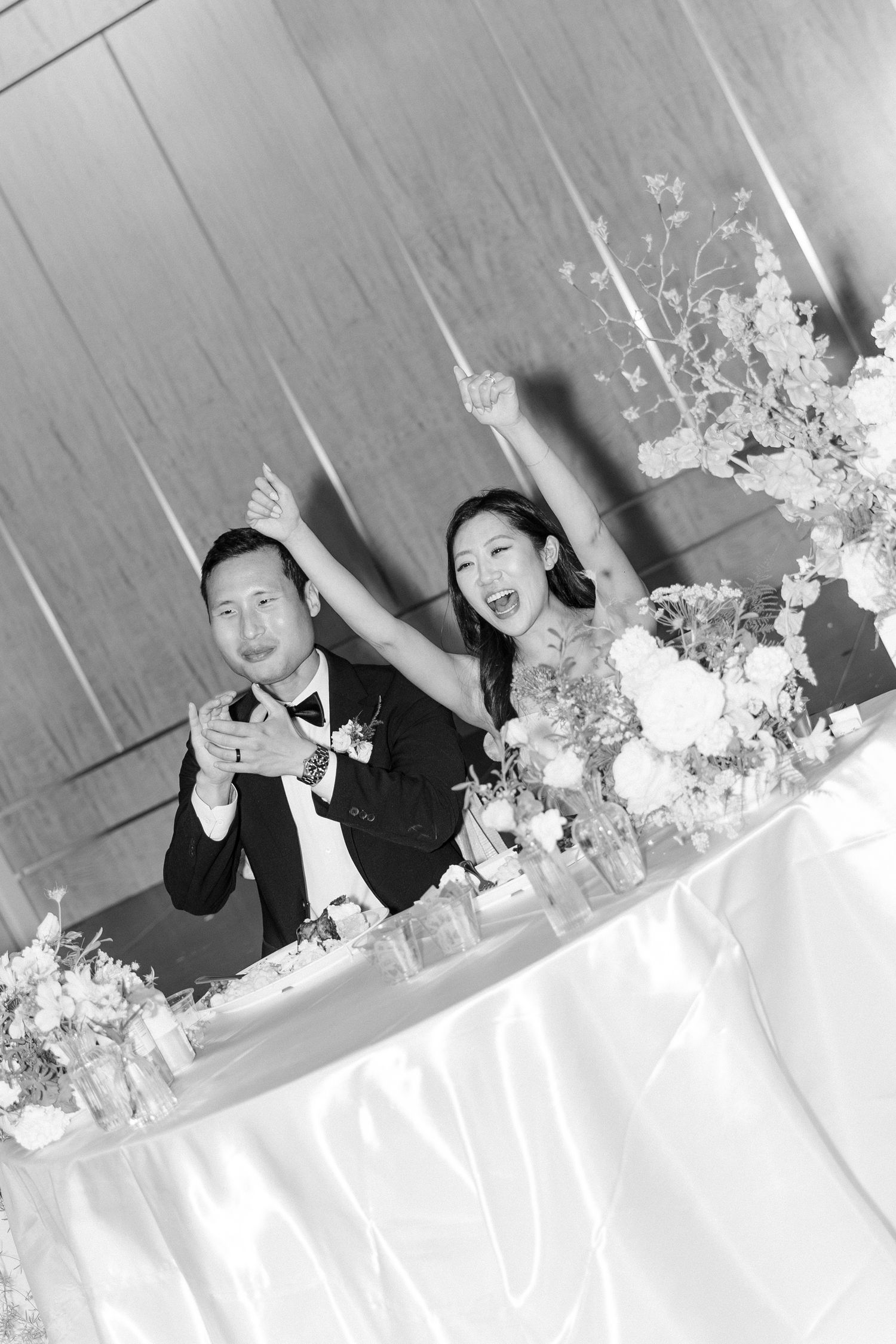 Black and white photo of a joyful bride and groom celebrating at their wedding reception table with floral arrangements.