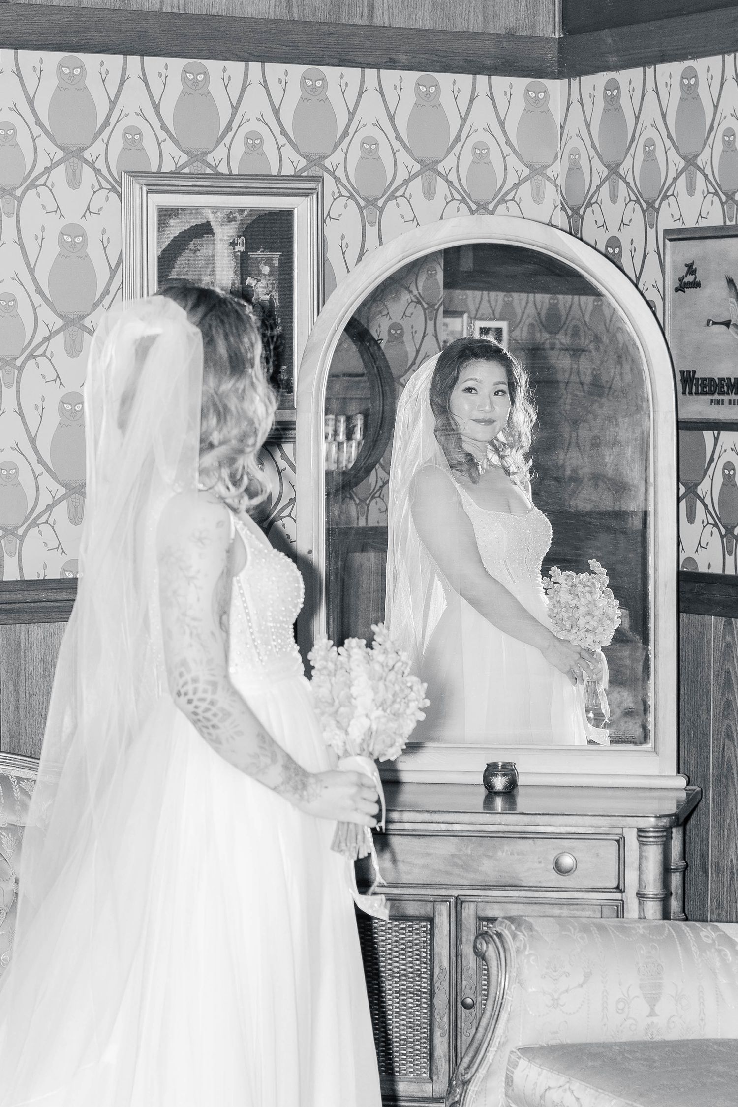Black and white photo of a bride in a lace gown holding a bouquet, gazing at her reflection in an ornate mirror.