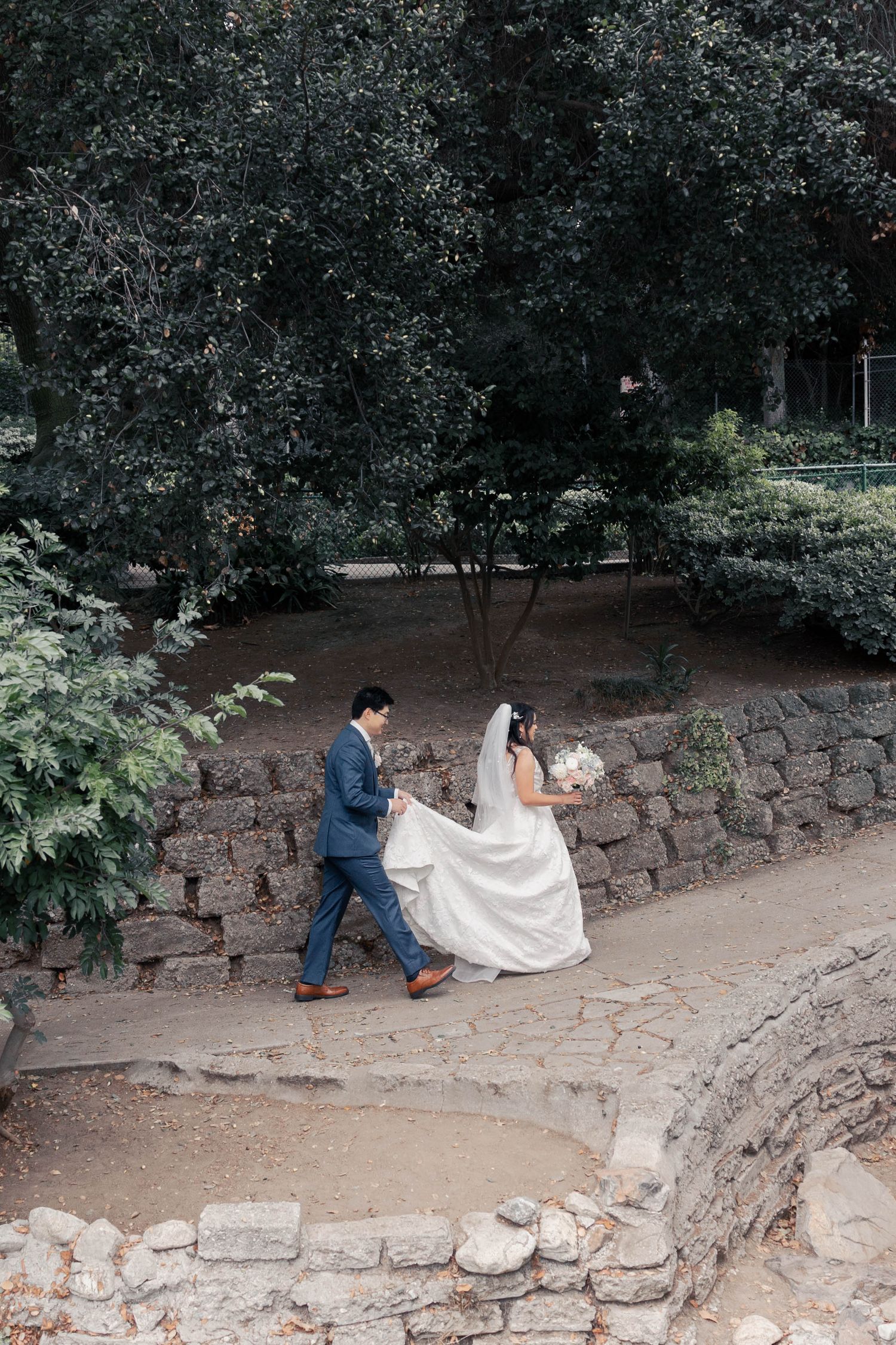 A couple in wedding attire walks along a rustic stone path in a garden setting.