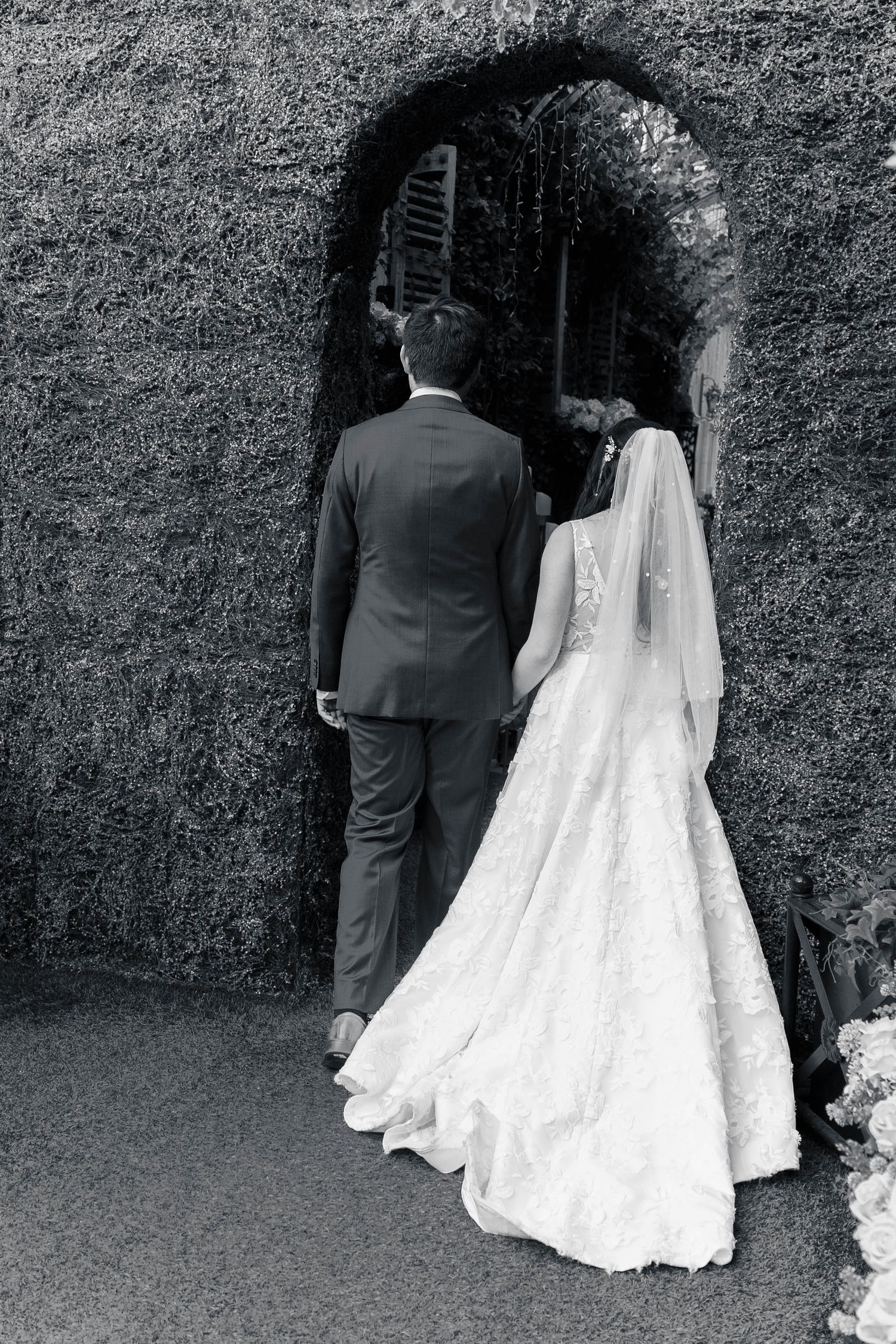 A bride and groom in formal attire stand before a stone archway entrance in a black and white wedding photograph.