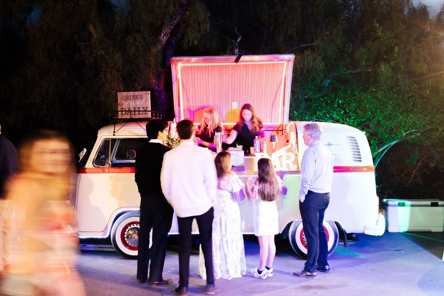Guests gather around a vintage VW bus food truck at an outdoor evening event with festive lighting.