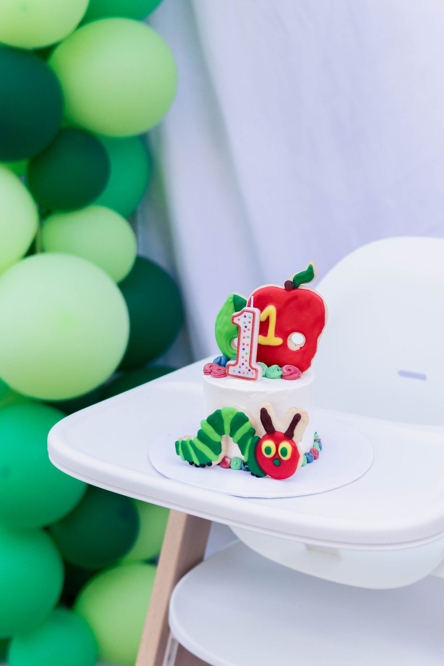 Decorative caterpillar-themed cake toppers displayed on white pedestals against a green balloon backdrop.