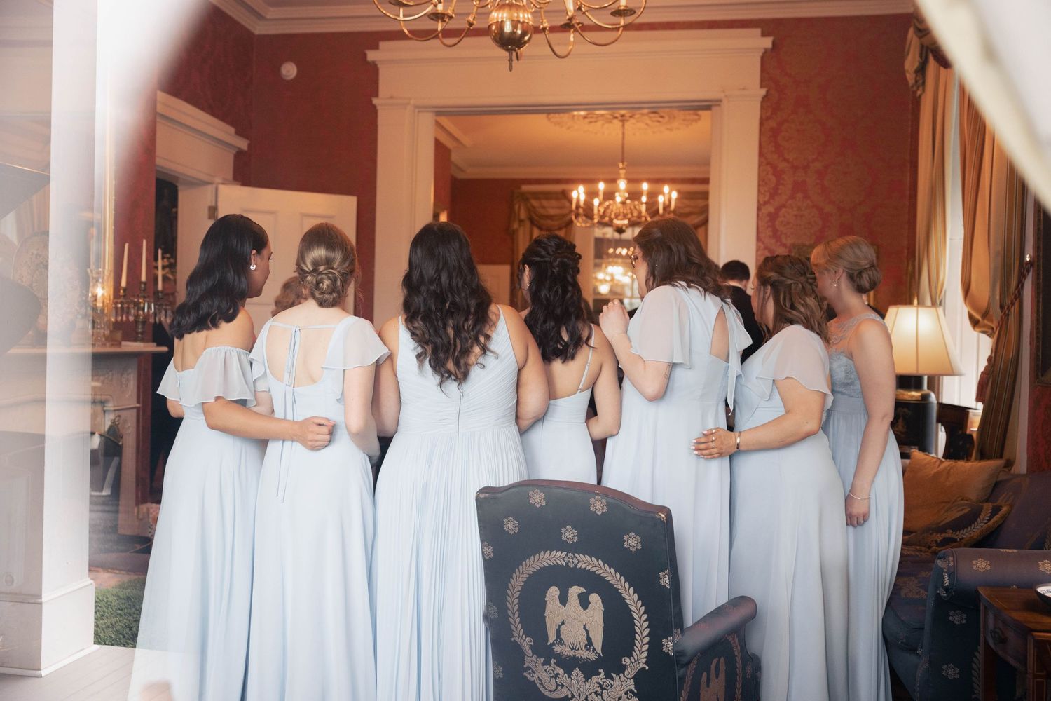 A group of bridesmaids in light blue dresses stand together in an elegant room with chandeliers.