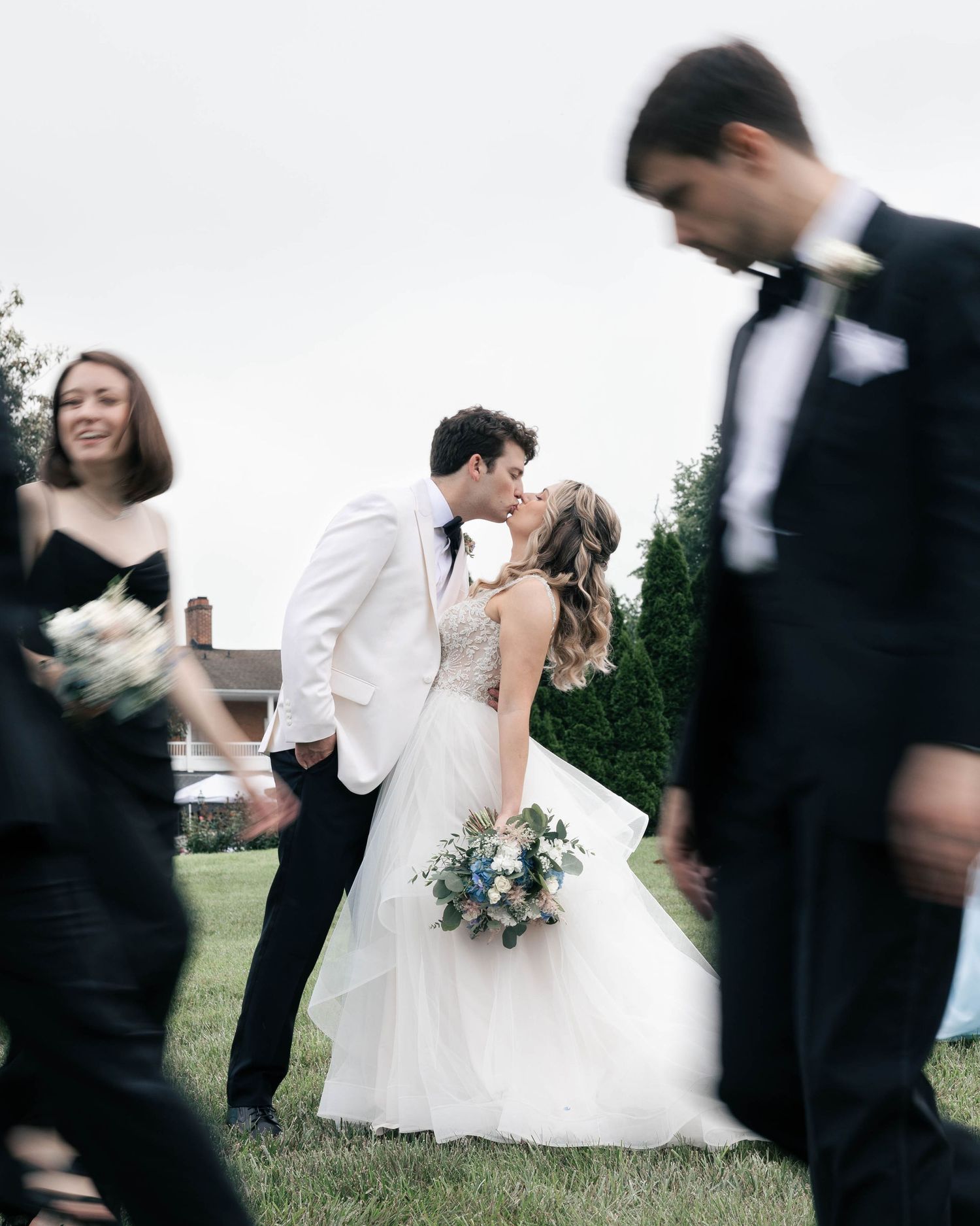 A romantic wedding moment captured in motion as guests walk past the couple sharing a kiss on a grassy lawn.