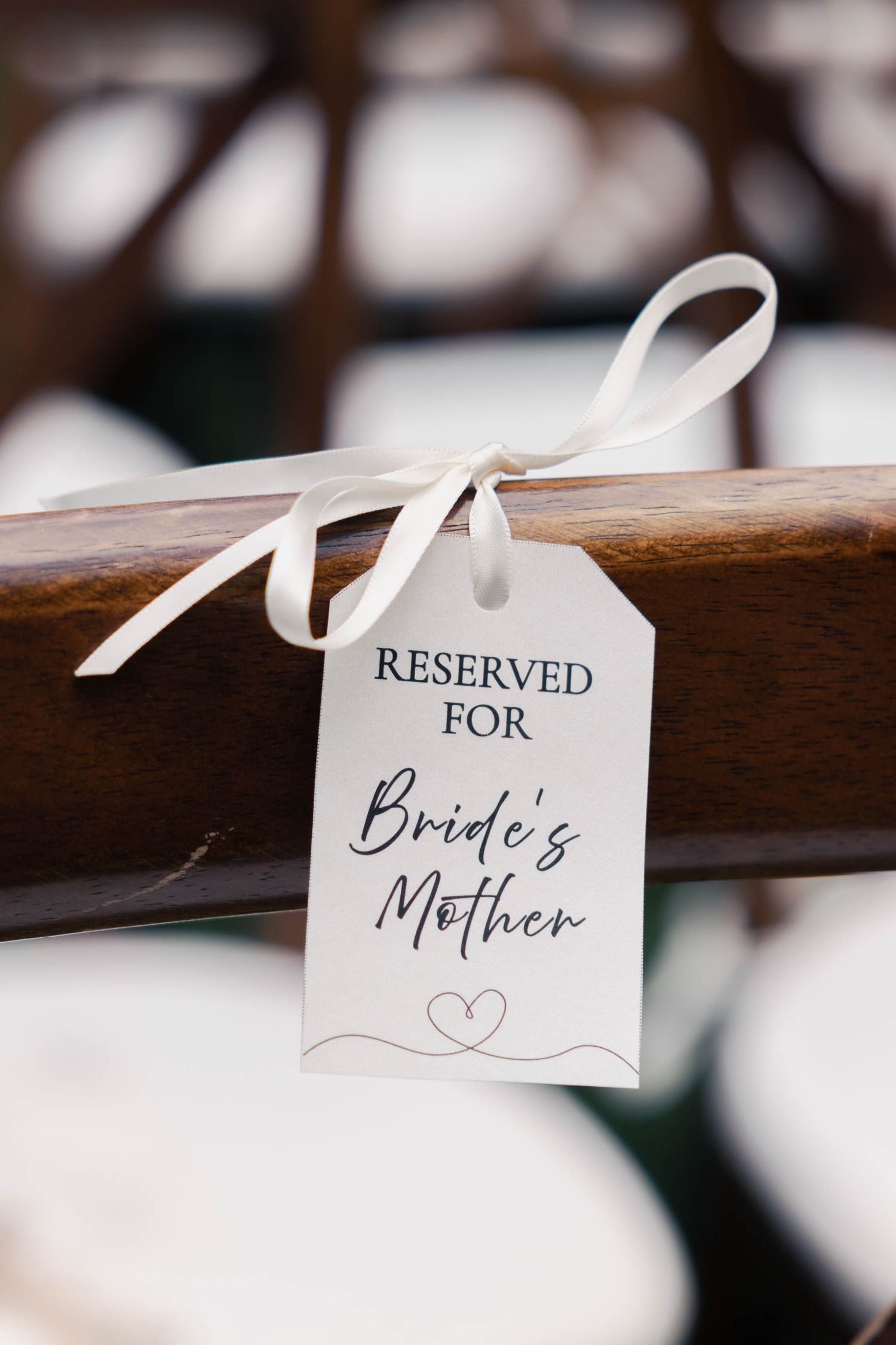A white tag with 'Reserved for Bride's Mother' text tied with a ribbon to a wooden chair at a wedding ceremony.