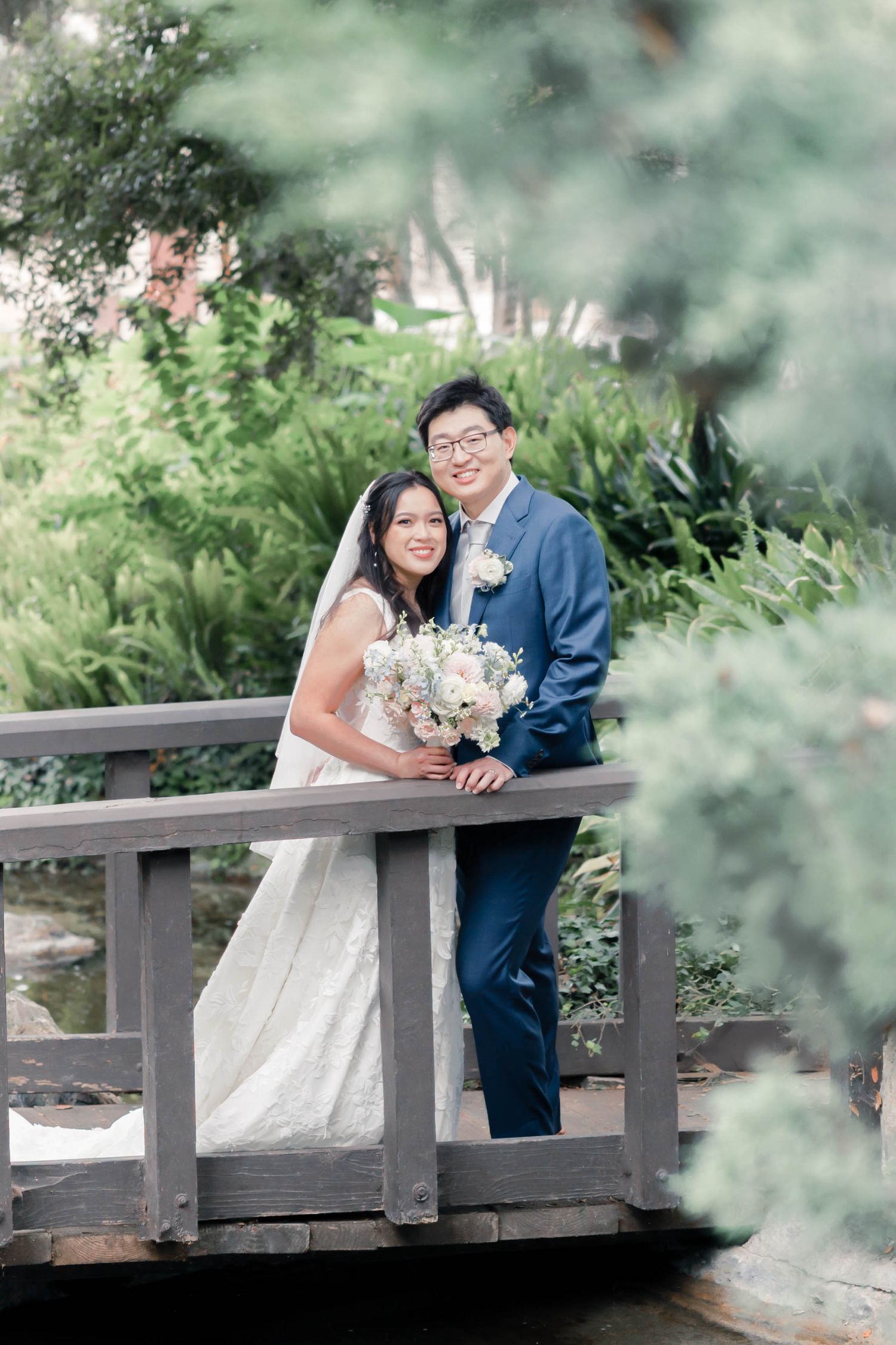 A couple in wedding attire poses together on a wooden bridge surrounded by lush greenery in a garden setting.