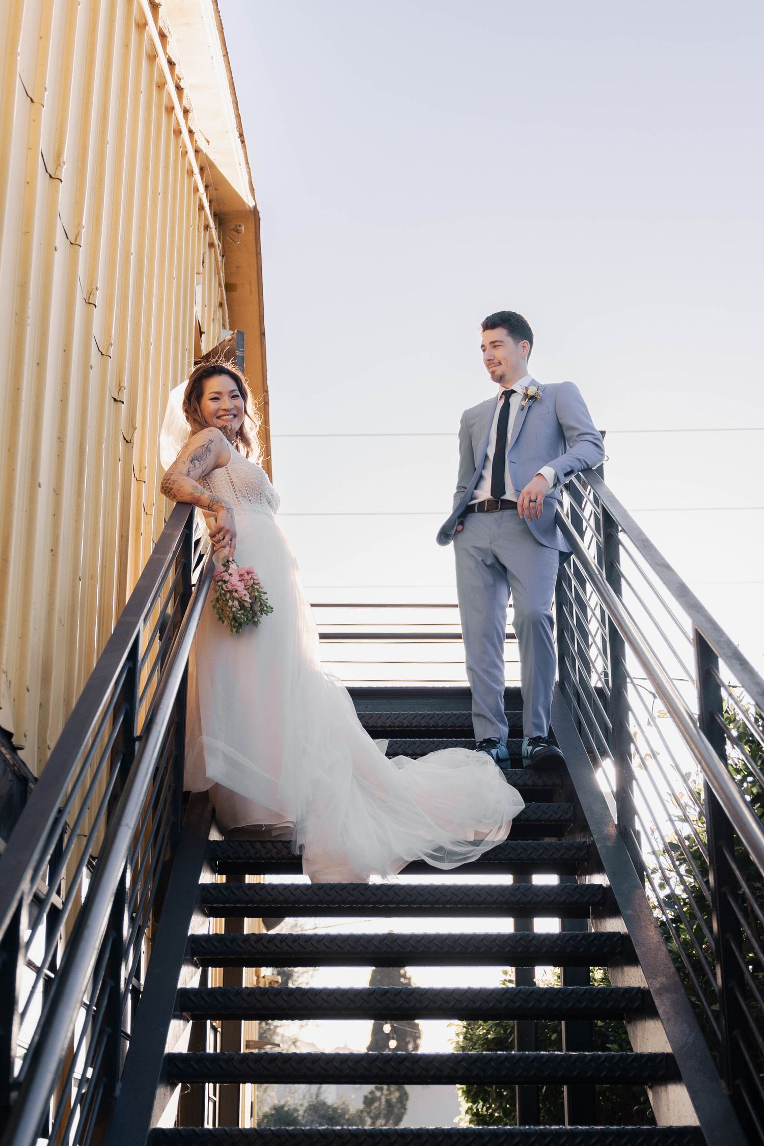 Wedding couple photographed in sequence descending modern metal stairs against a bright sky.