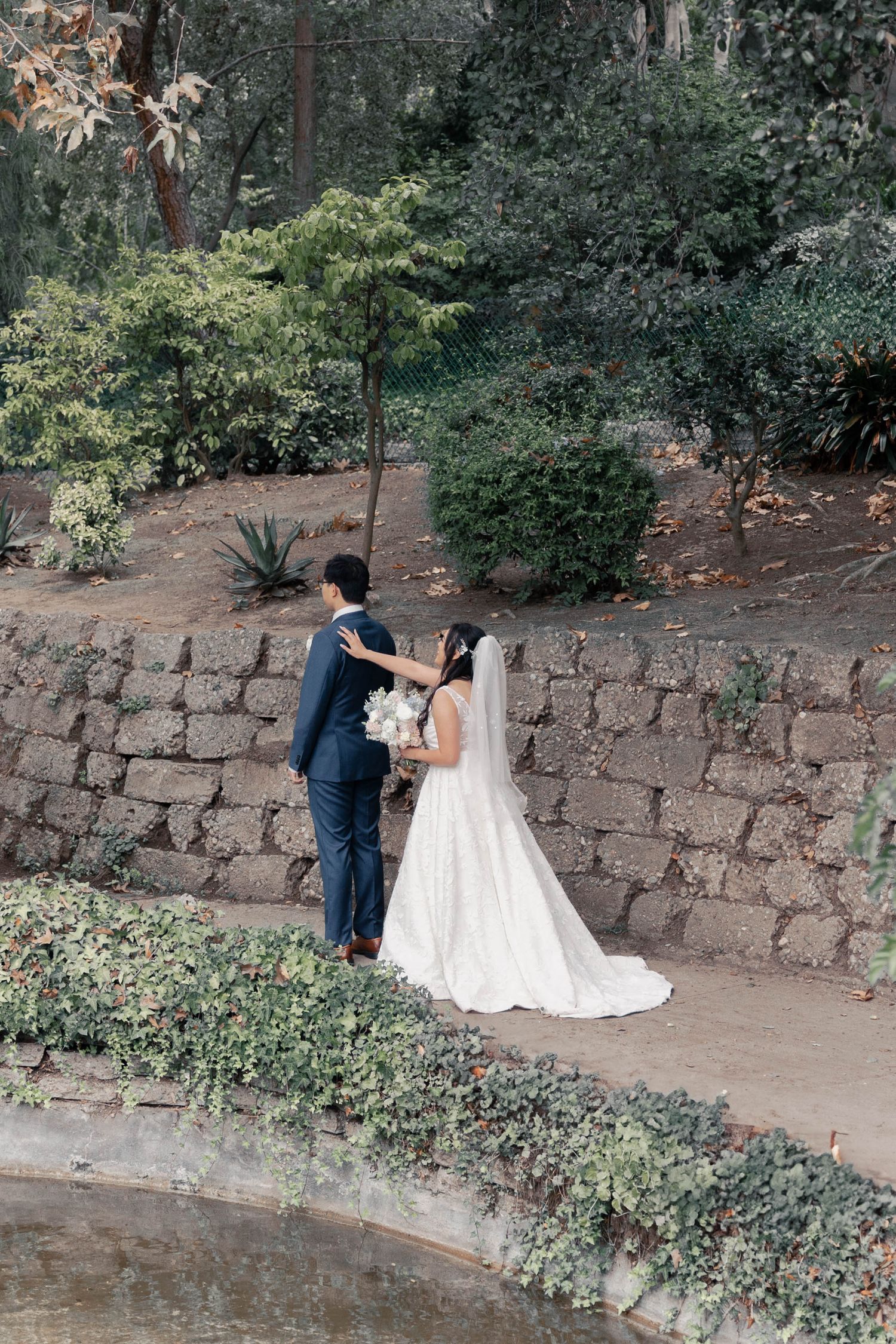 A couple in wedding attire dance together on a stone-walled garden path surrounded by lush greenery.