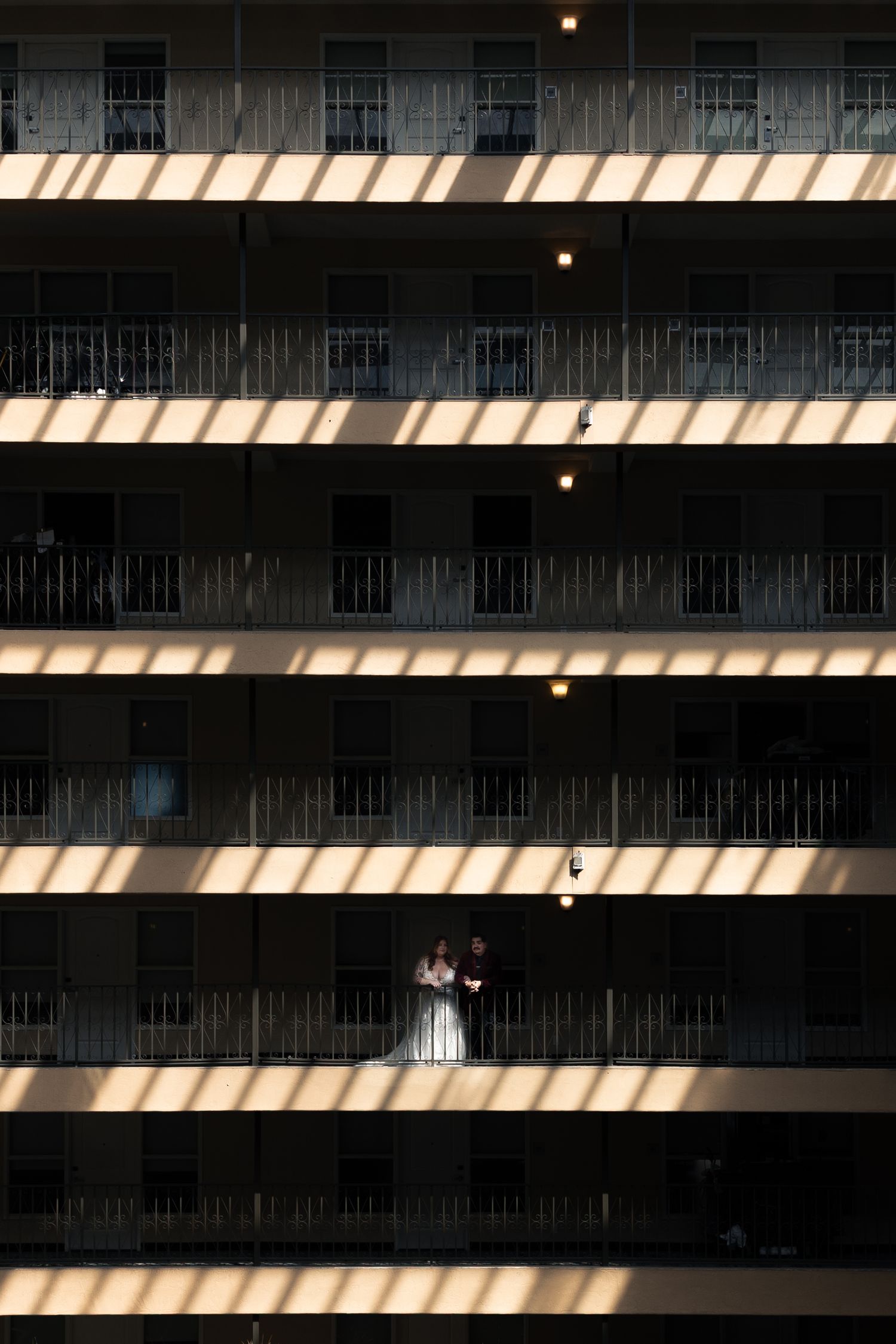 Bride and groom standing on a sunlit hotel balcony, surrounded by rows of shadowed floors in a tall building.
