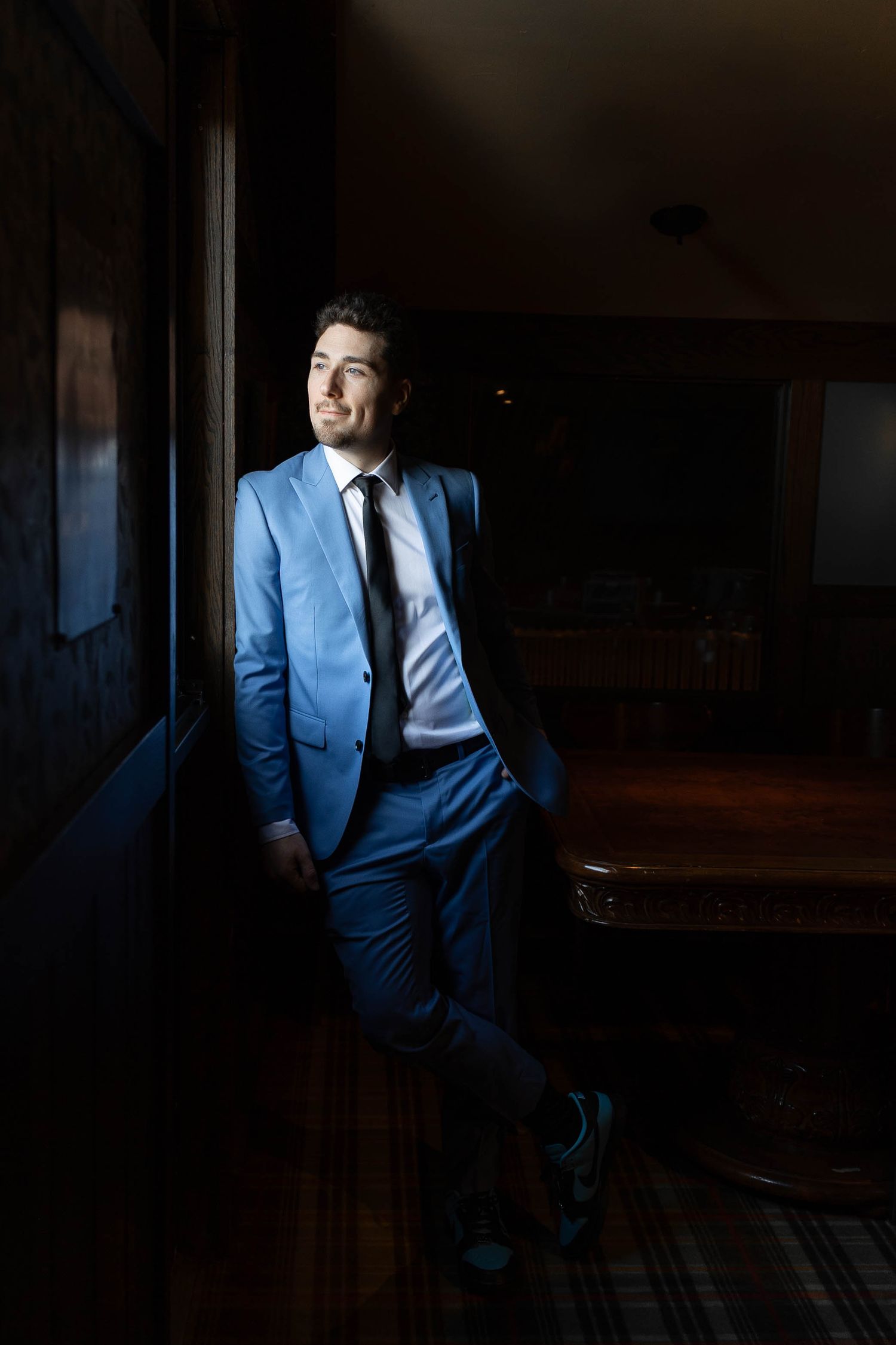 A figure in a blue suit sits in a dark room illuminated by dramatic window lighting.