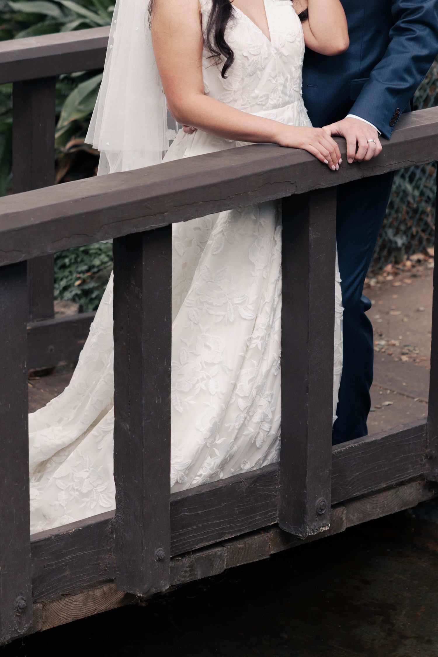 Close-up view of a bride in white lace dress and groom in navy suit standing together on a wooden bridge.