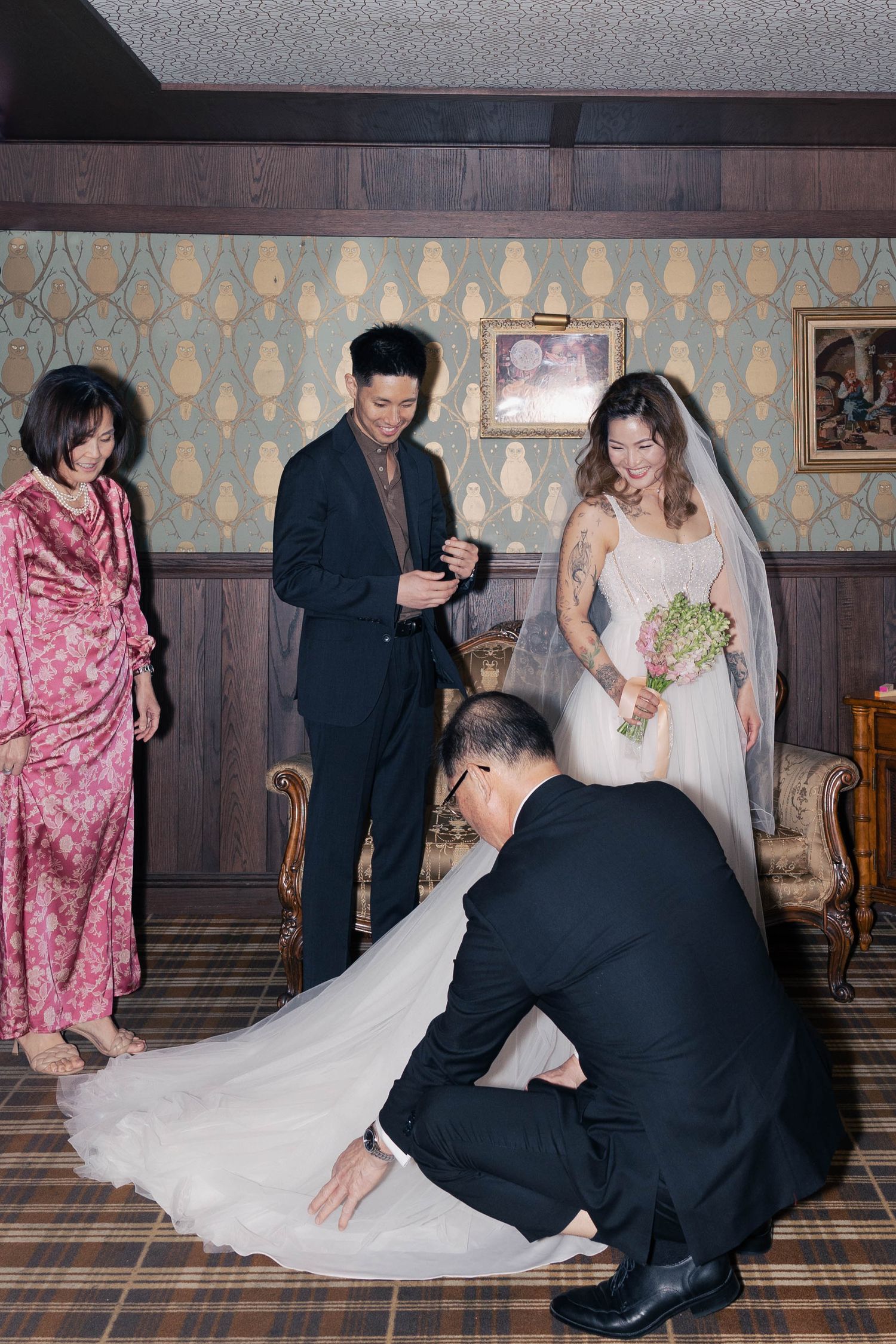 Wedding party helps adjust the bride's gown train in a vintage-styled room with retro wallpaper and wood paneling.