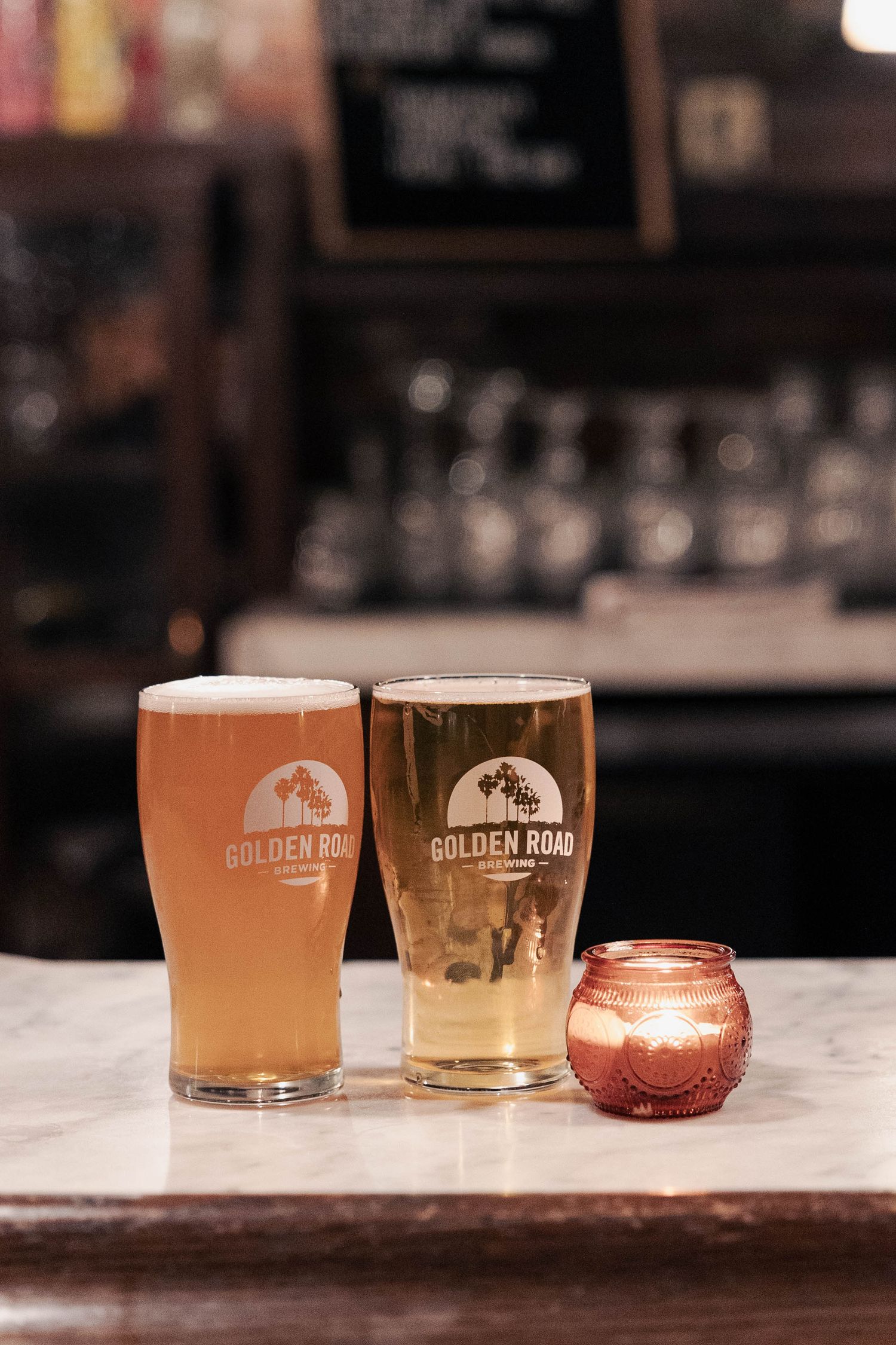 Two glasses of craft beer from Golden Road Brewing sit on a marble bar counter with a small candle between them.